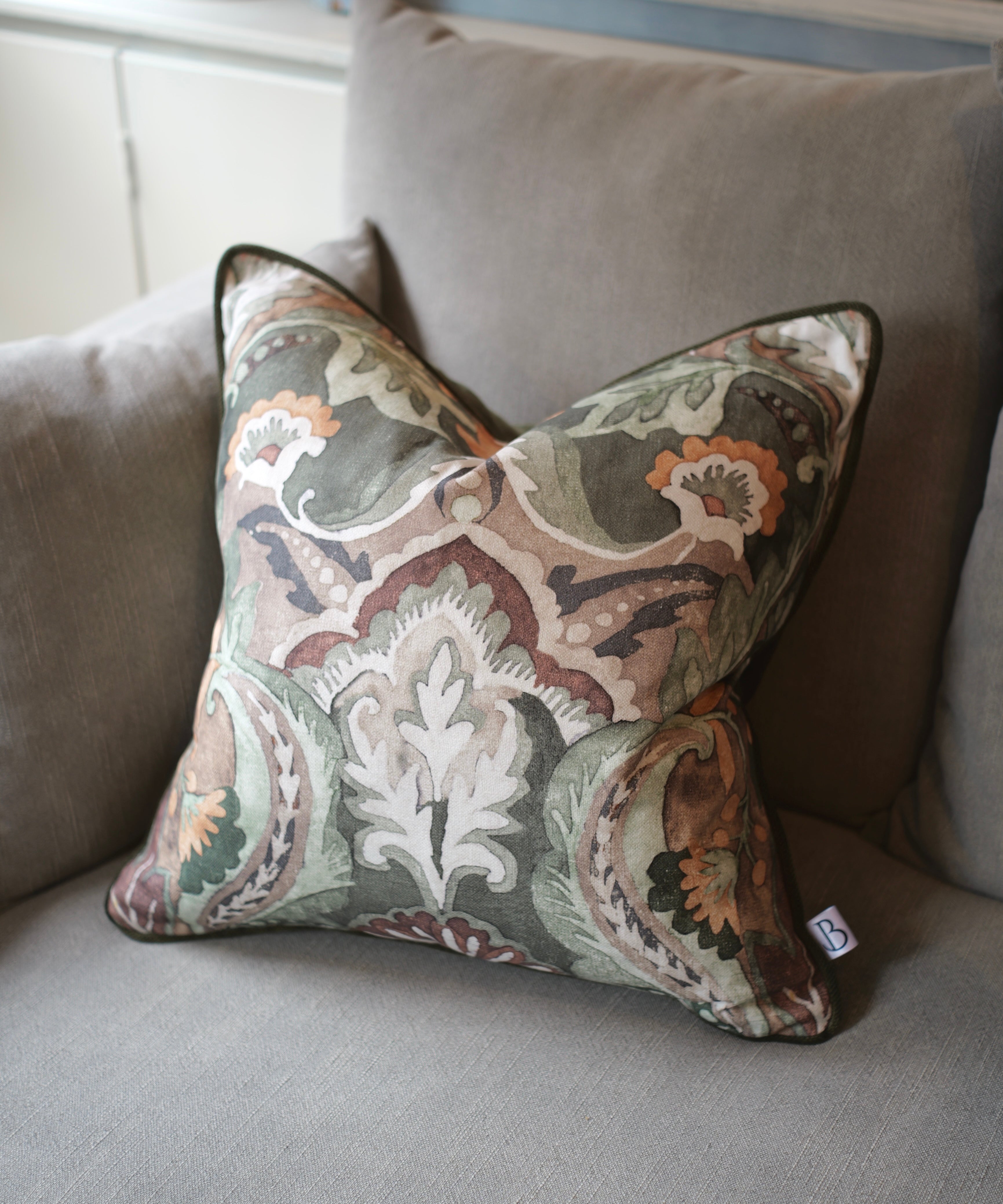A square decorative throw pillow with an intricate paisley and floral damask pattern in muted tones of green, beige, brown, and orange, resting on a gray fabric sofa.