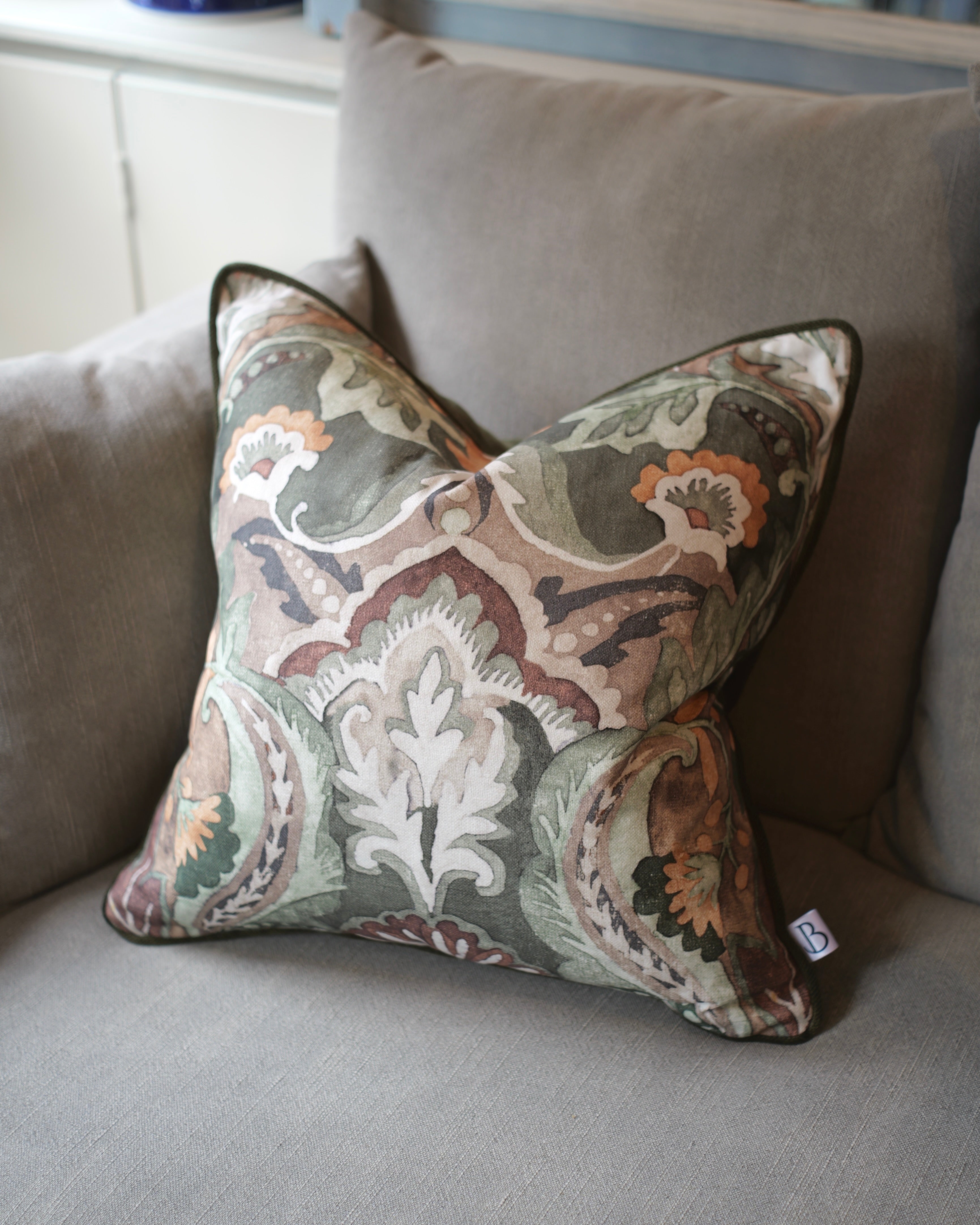 A square decorative throw pillow with an intricate paisley and floral damask pattern in muted tones of green, beige, brown, and orange, resting on a gray fabric sofa.