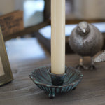 A close-up of a rustic wooden table styled with a textured teal metal candle holder, a cream pillar candle, and two metallic silver bird figurines.