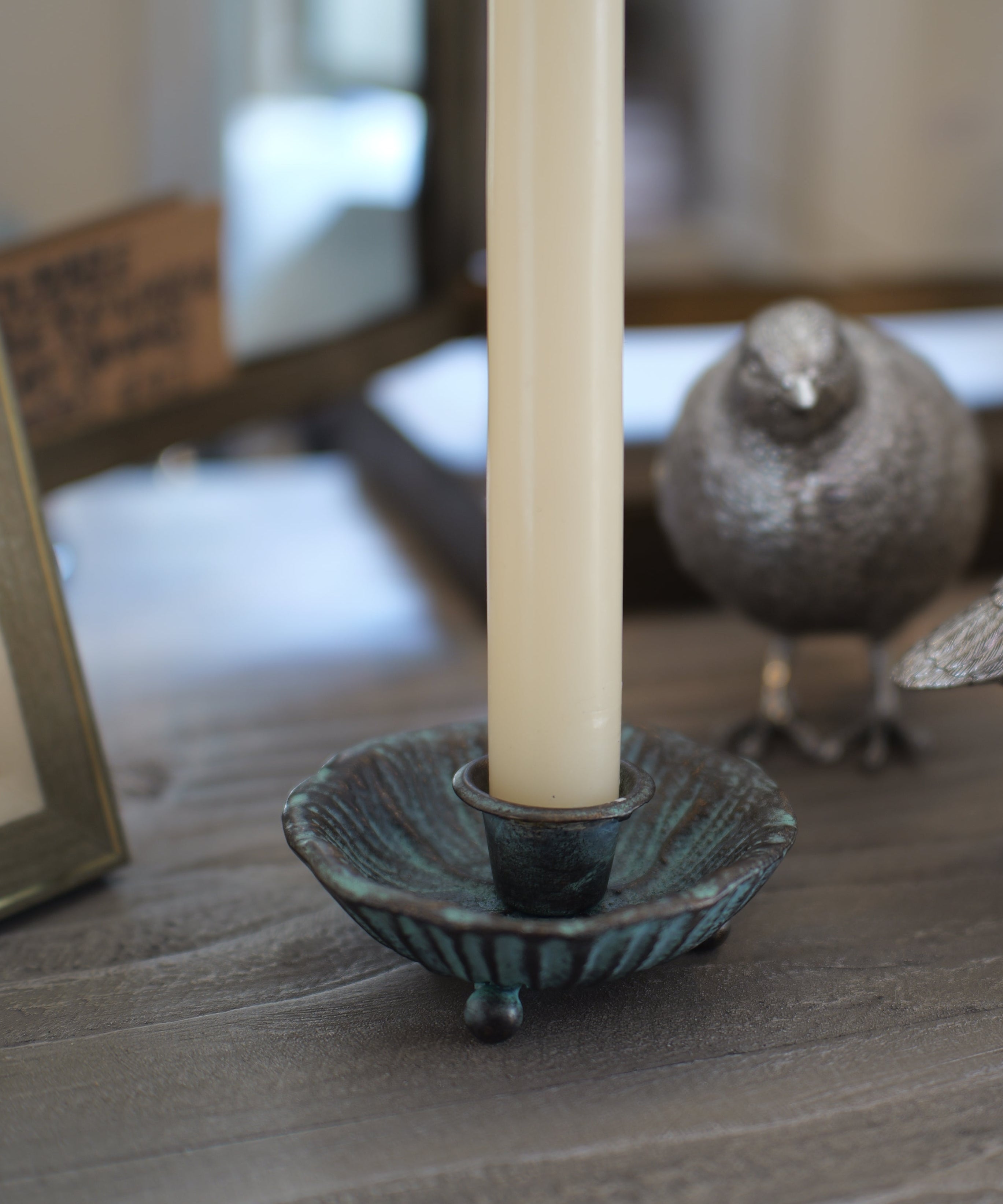 A close-up of a rustic wooden table styled with a textured teal metal candle holder, a cream pillar candle, and two metallic silver bird figurines.