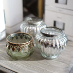 Three decorative glass candle holders with metallic trim, including one green-tinted glass holder with bronze detailing, one silver-tinted glass holder, and one clear glass holder with silver trim, arranged on a rustic wooden surface.