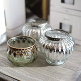 Three decorative glass candle holders with metallic trim, including one green-tinted glass holder with bronze detailing, one silver-tinted glass holder, and one clear glass holder with silver trim, arranged on a rustic wooden surface.