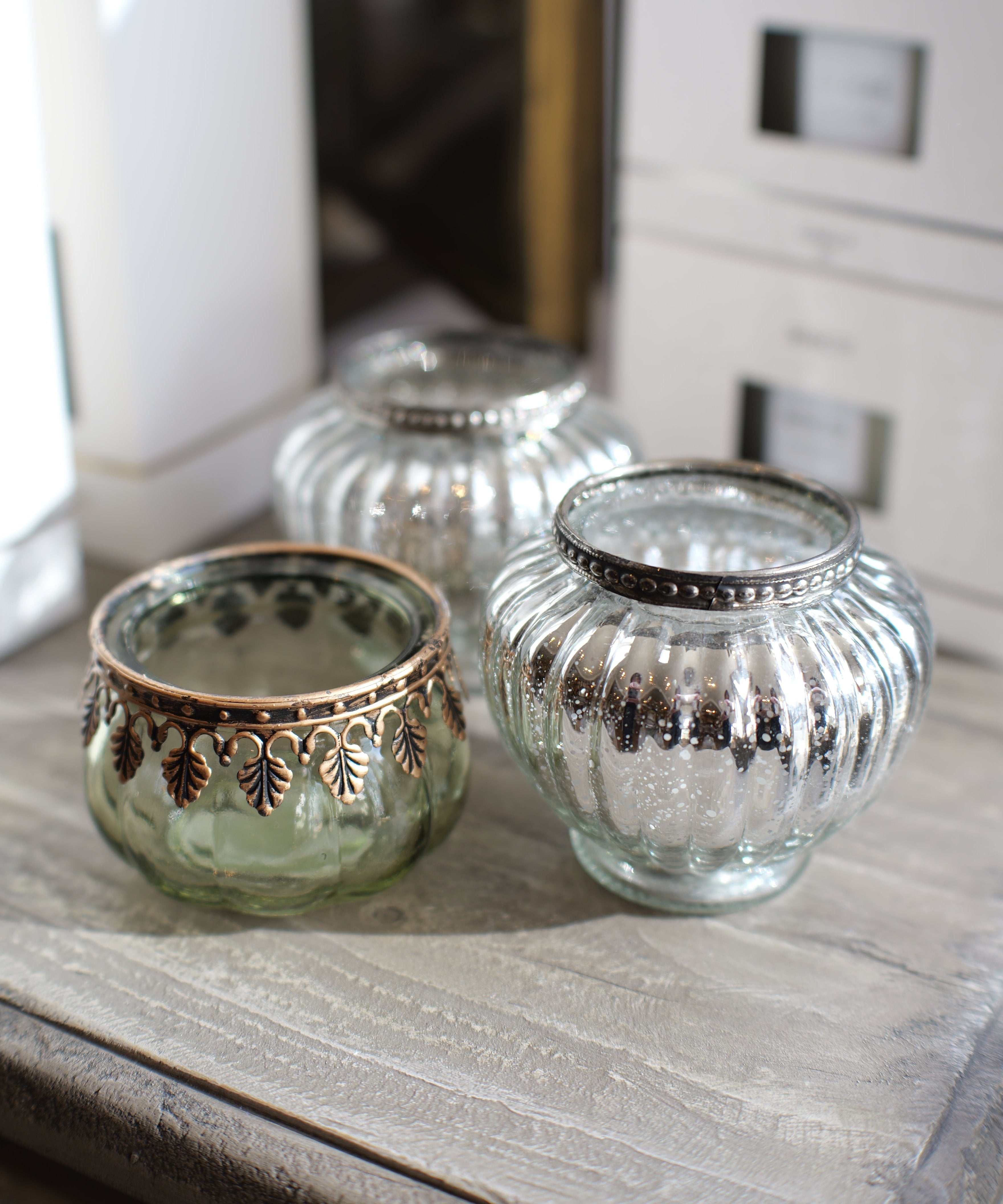 Three decorative glass candle holders with metallic trim, including one green-tinted glass holder with bronze detailing, one silver-tinted glass holder, and one clear glass holder with silver trim, arranged on a rustic wooden surface.