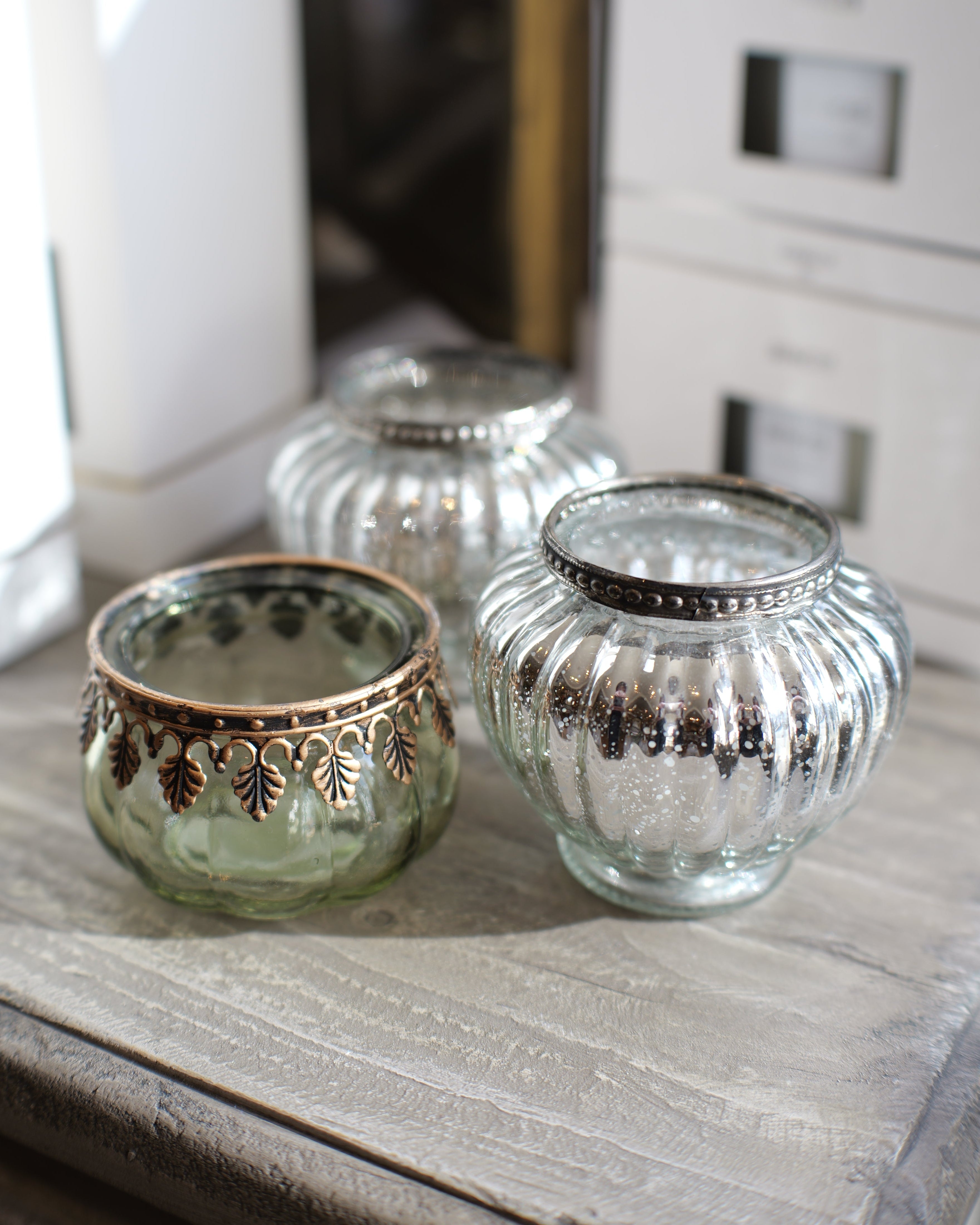 Three decorative glass candle holders with metallic trim, including one green-tinted glass holder with bronze detailing, one silver-tinted glass holder, and one clear glass holder with silver trim, arranged on a rustic wooden surface.