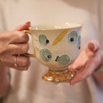 A person holding a ceramic teacup with a gold base and handle, decorated with blue-gray and gold abstract spots on a cream-colored body.