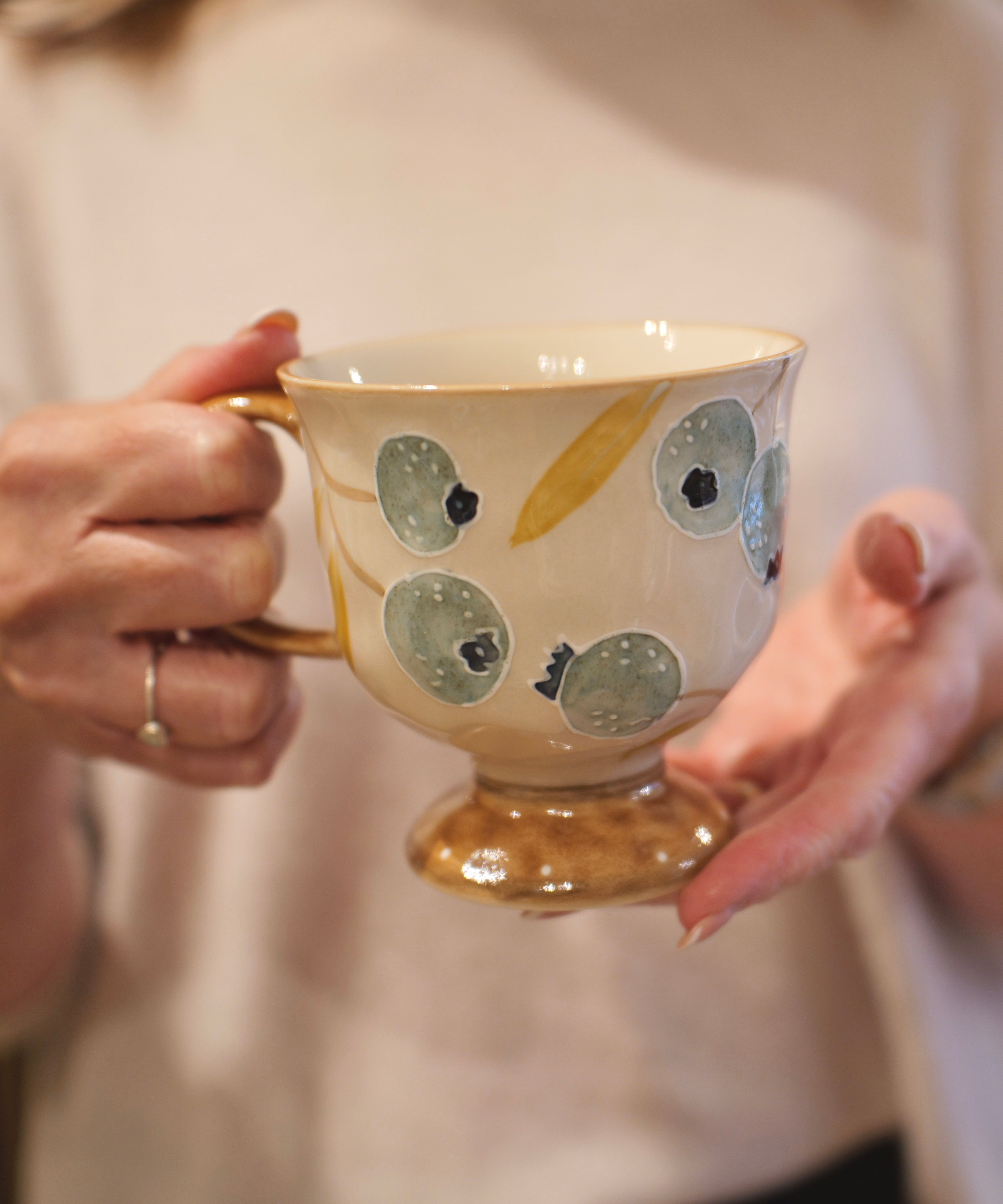 A person holding a ceramic teacup with a gold base and handle, decorated with blue-gray and gold abstract spots on a cream-colored body.