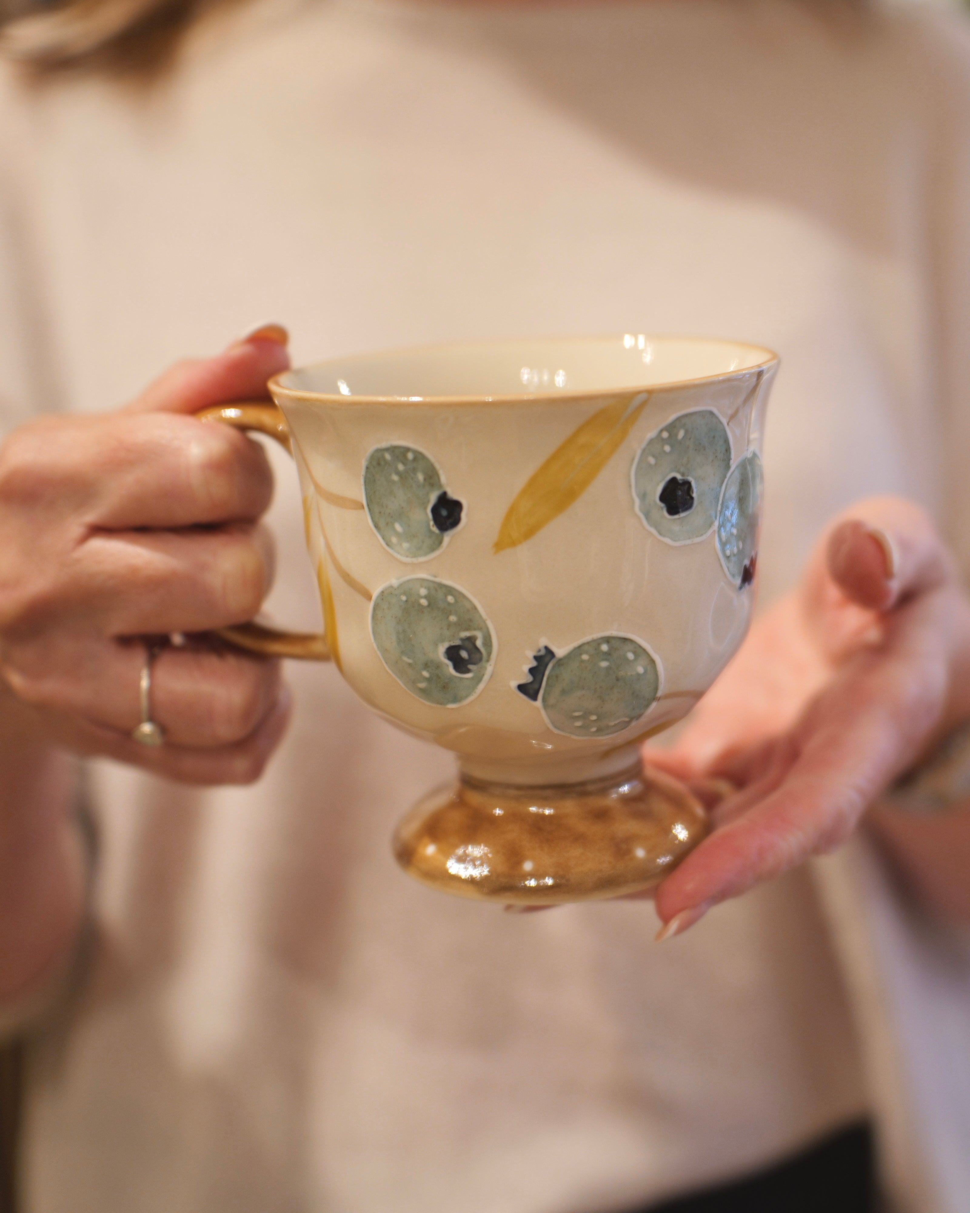 A person holding a ceramic teacup with a gold base and handle, decorated with blue-gray and gold abstract spots on a cream-colored body.