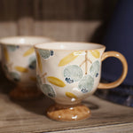 A ceramic coffee mug with a footed base, decorated with a pattern of blueberries and leaves in muted blue, yellow, and beige tones, sitting on a rustic wooden shelf next to another blurred mug.