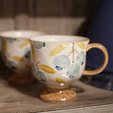 A ceramic coffee mug with a footed base, decorated with a pattern of blueberries and leaves in muted blue, yellow, and beige tones, sitting on a rustic wooden shelf next to another blurred mug.