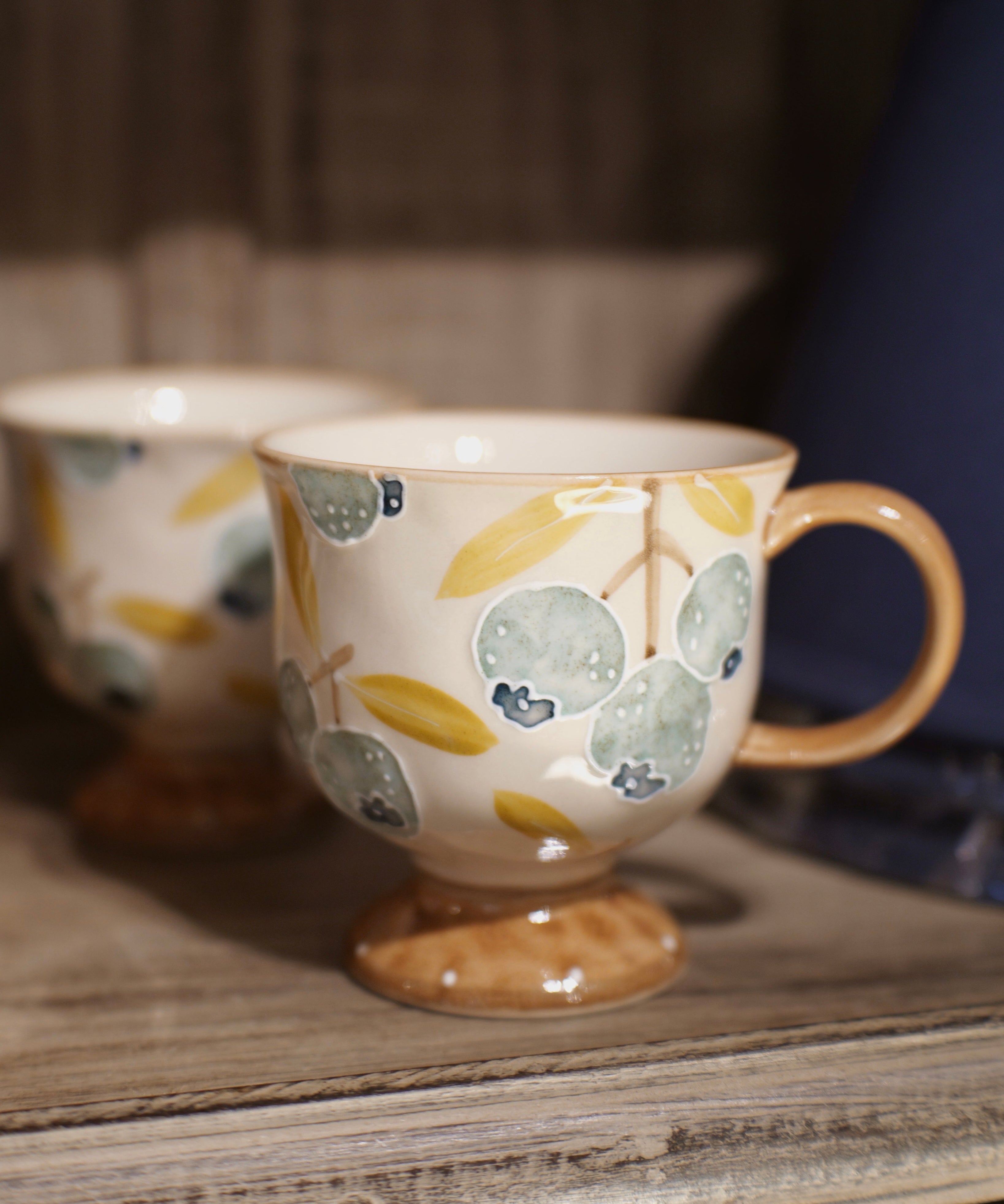 A ceramic coffee mug with a footed base, decorated with a pattern of blueberries and leaves in muted blue, yellow, and beige tones, sitting on a rustic wooden shelf next to another blurred mug.