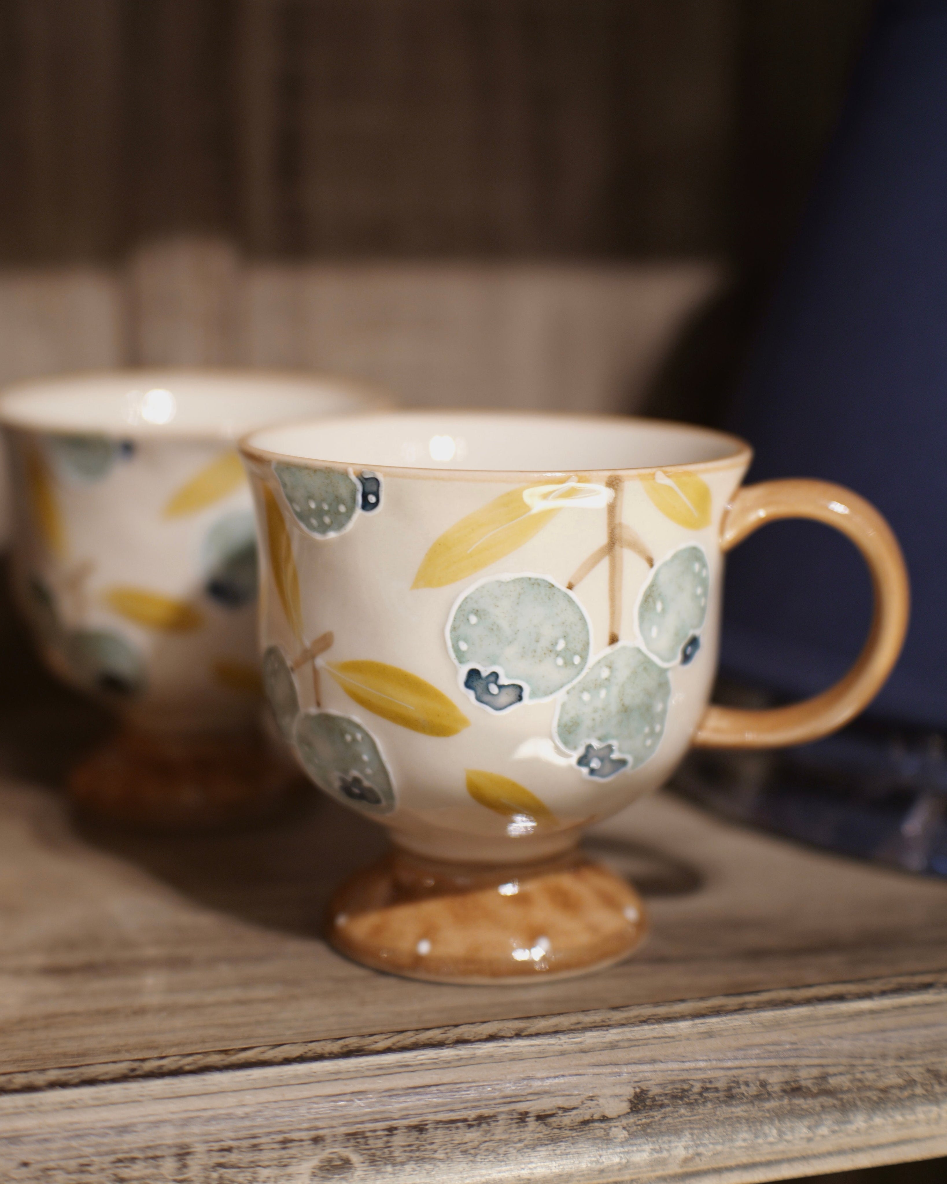 A ceramic coffee mug with a footed base, decorated with a pattern of blueberries and leaves in muted blue, yellow, and beige tones, sitting on a rustic wooden shelf next to another blurred mug.
