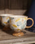 A ceramic coffee mug with a footed base, decorated with a pattern of blueberries and leaves in muted blue, yellow, and beige tones, sitting on a rustic wooden shelf next to another blurred mug.
