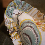 A close-up of a decorative throw pillow with a ruffled edge, featuring a cream background and a teal, brown, and mustard yellow paisley pattern, resting on a dark gray couch.