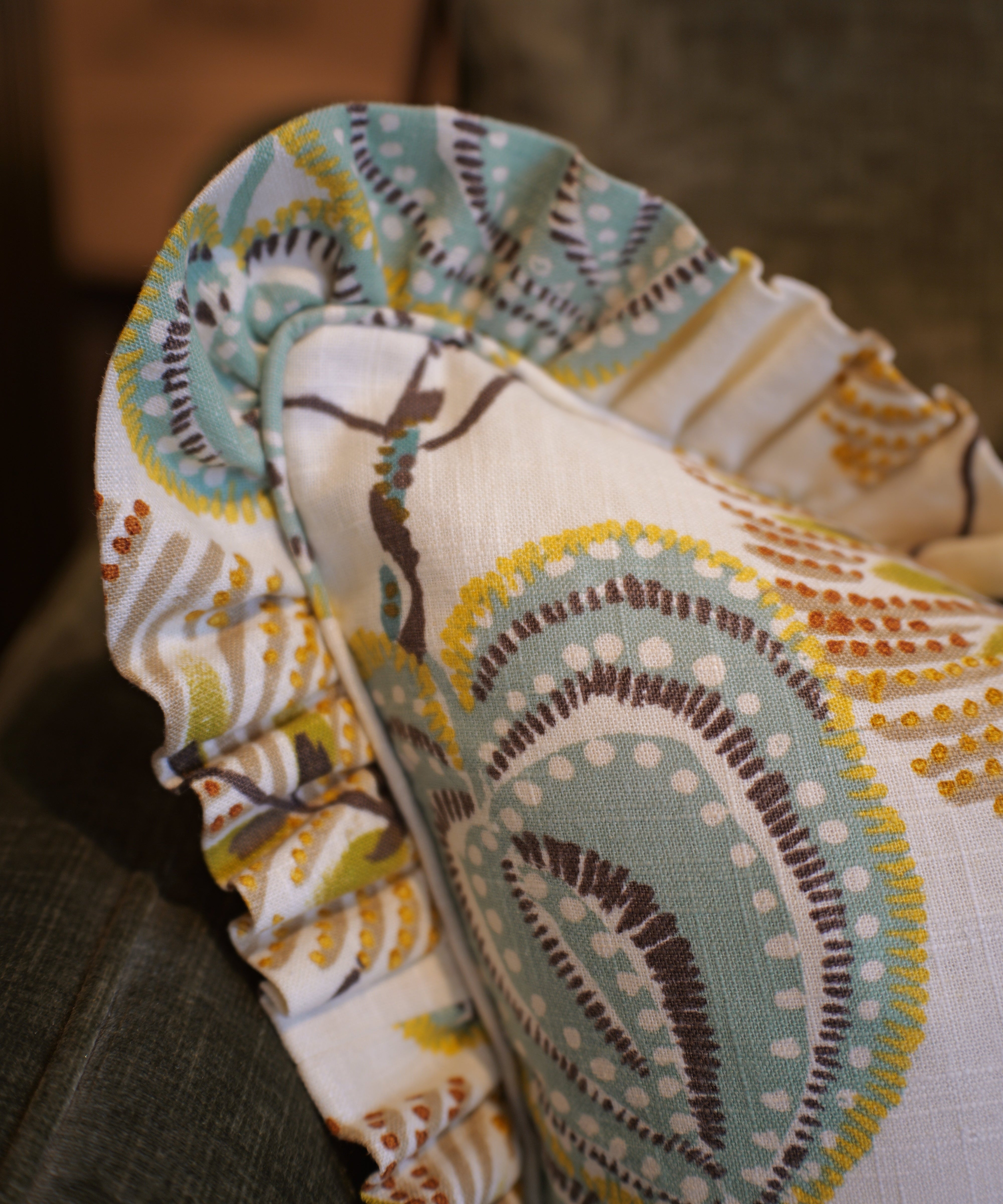 A close-up of a decorative throw pillow with a ruffled edge, featuring a cream background and a teal, brown, and mustard yellow paisley pattern, resting on a dark gray couch.