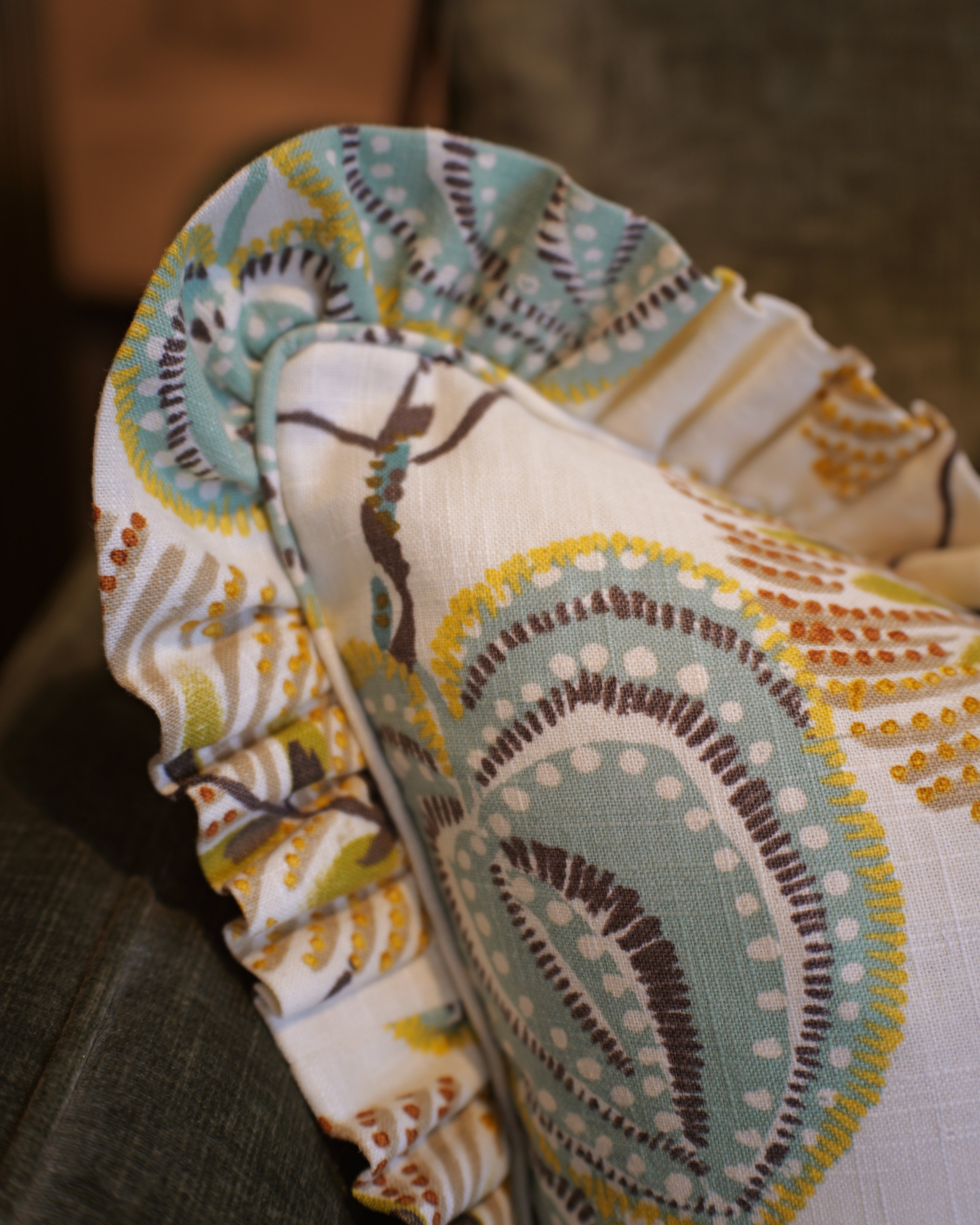A close-up of a decorative throw pillow with a ruffled edge, featuring a cream background and a teal, brown, and mustard yellow paisley pattern, resting on a dark gray couch.