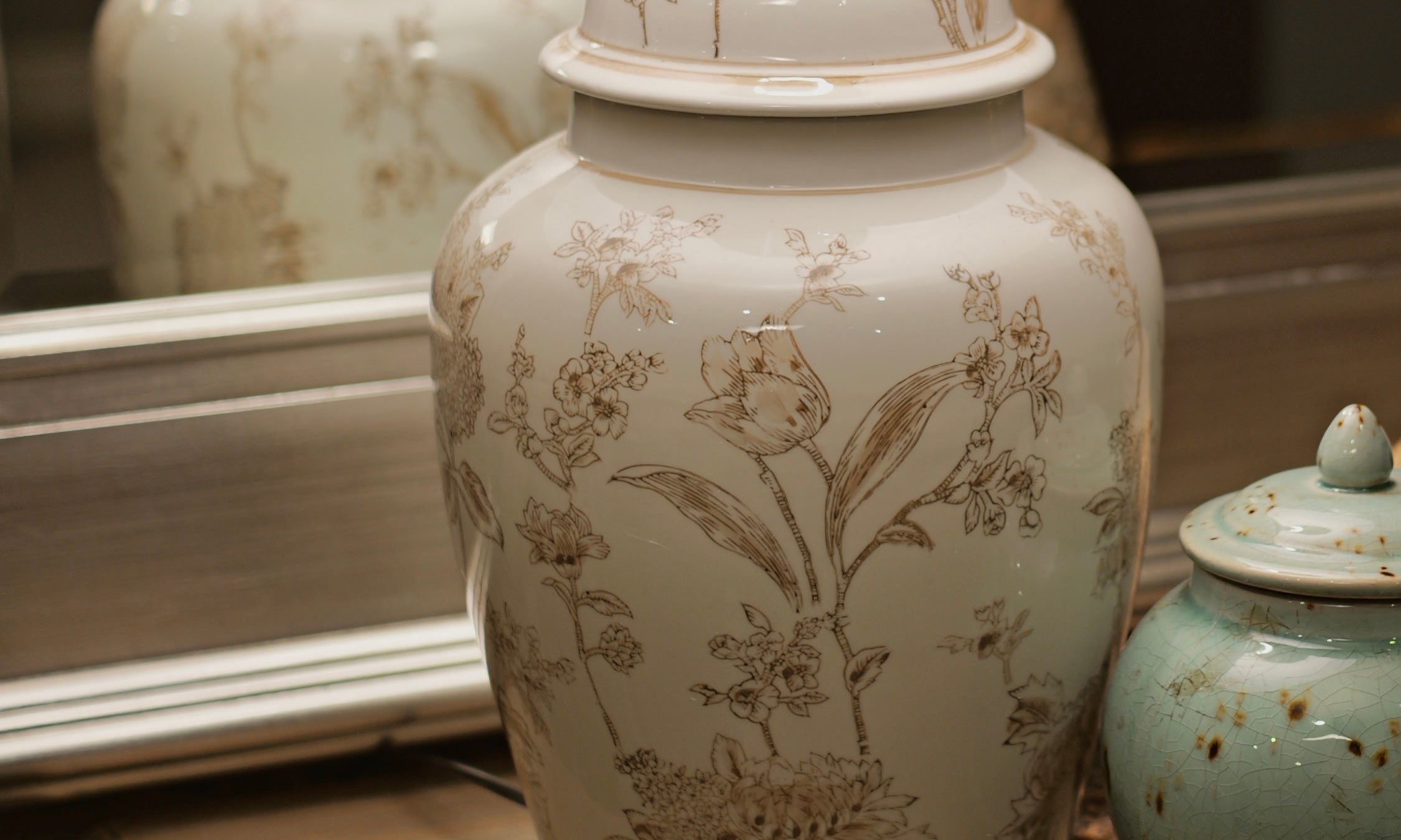 A vintage-style table lamp with a large cream fabric drum shade and a white ceramic jar base decorated with delicate beige botanical illustrations, displayed on a rustic wooden dresser alongside a small speckled teal jar and a decorative bird figurine.