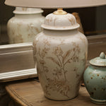 A vintage-style table lamp with a large cream fabric drum shade and a white ceramic jar base decorated with delicate beige botanical illustrations, displayed on a rustic wooden dresser alongside a small speckled teal jar and a decorative bird figurine.
