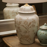A vintage-style table lamp with a large cream fabric drum shade and a white ceramic jar base decorated with delicate beige botanical illustrations, displayed on a rustic wooden dresser alongside a small speckled teal jar and a decorative bird figurine.