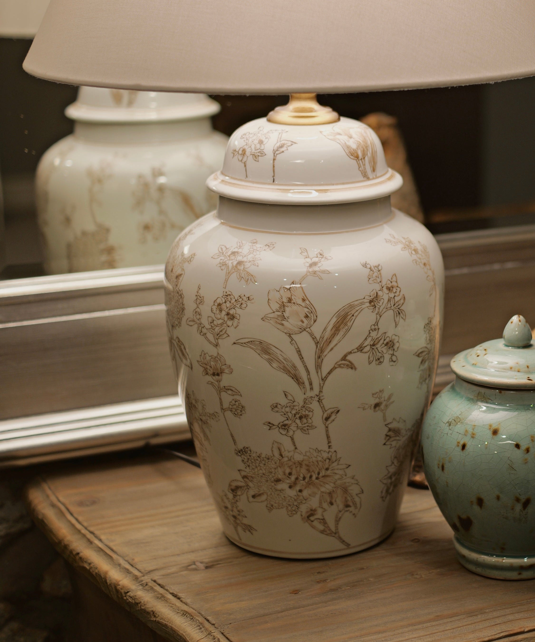 A vintage-style table lamp with a large cream fabric drum shade and a white ceramic jar base decorated with delicate beige botanical illustrations, displayed on a rustic wooden dresser alongside a small speckled teal jar and a decorative bird figurine.
