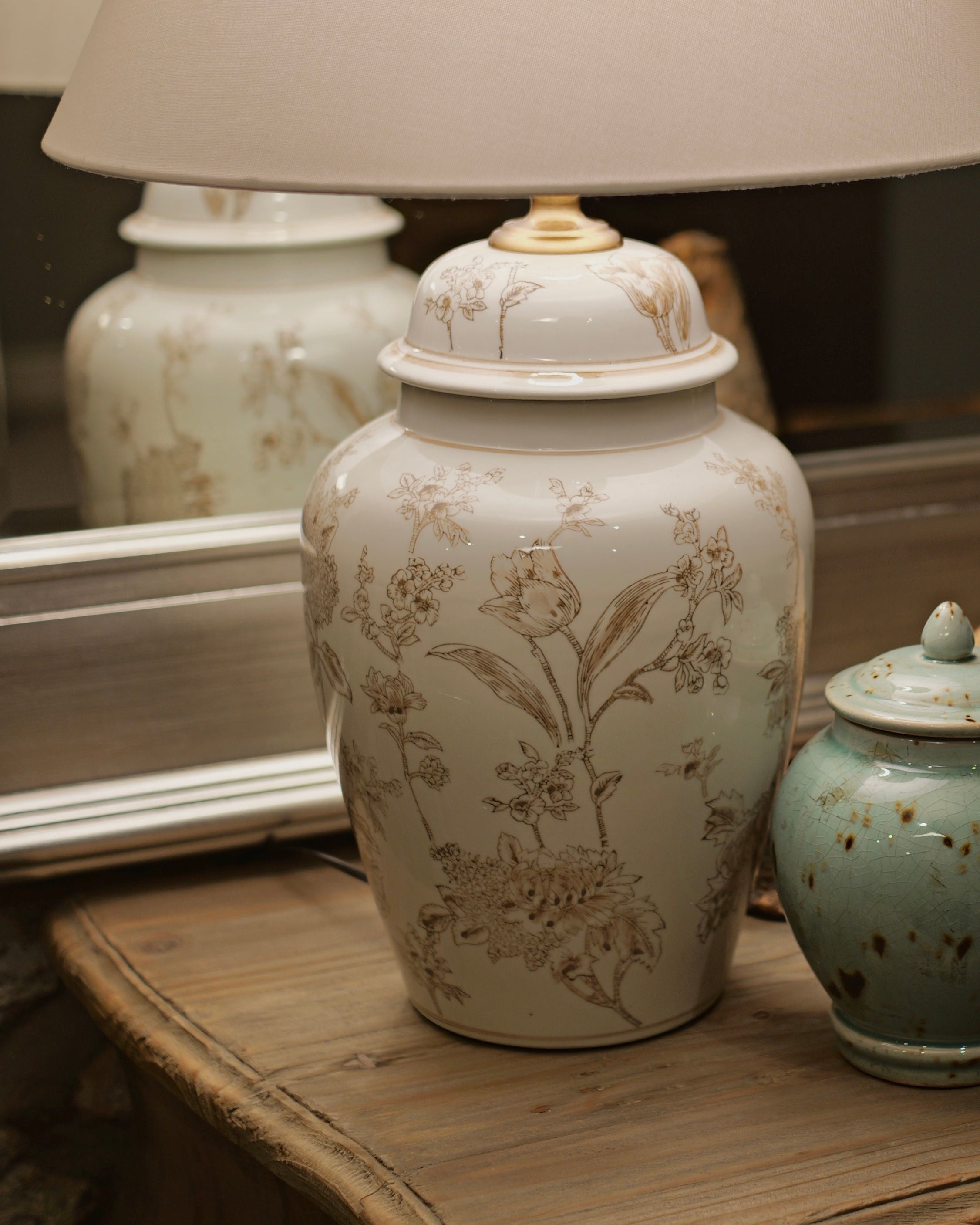 A vintage-style table lamp with a large cream fabric drum shade and a white ceramic jar base decorated with delicate beige botanical illustrations, displayed on a rustic wooden dresser alongside a small speckled teal jar and a decorative bird figurine.