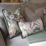 Two decorative throw pillows with a muted paisley pattern in sage green, dusty pink, and cream, styled on a gray sofa with a striped knit blanket.