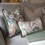 Two decorative throw pillows with a muted paisley pattern in sage green, dusty pink, and cream, styled on a gray sofa with a striped knit blanket.