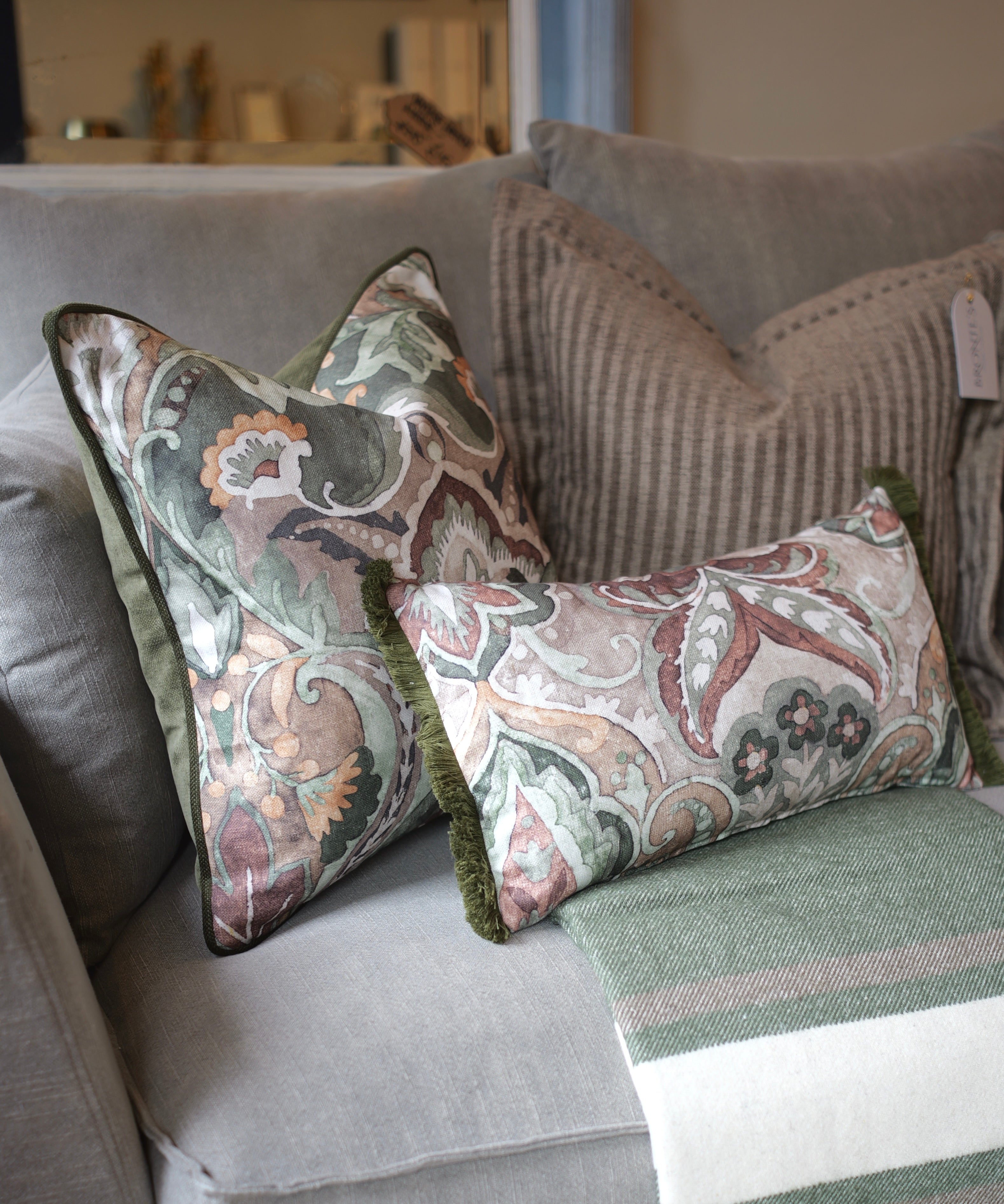 Two decorative throw pillows with a muted paisley pattern in sage green, dusty pink, and cream, styled on a gray sofa with a striped knit blanket.