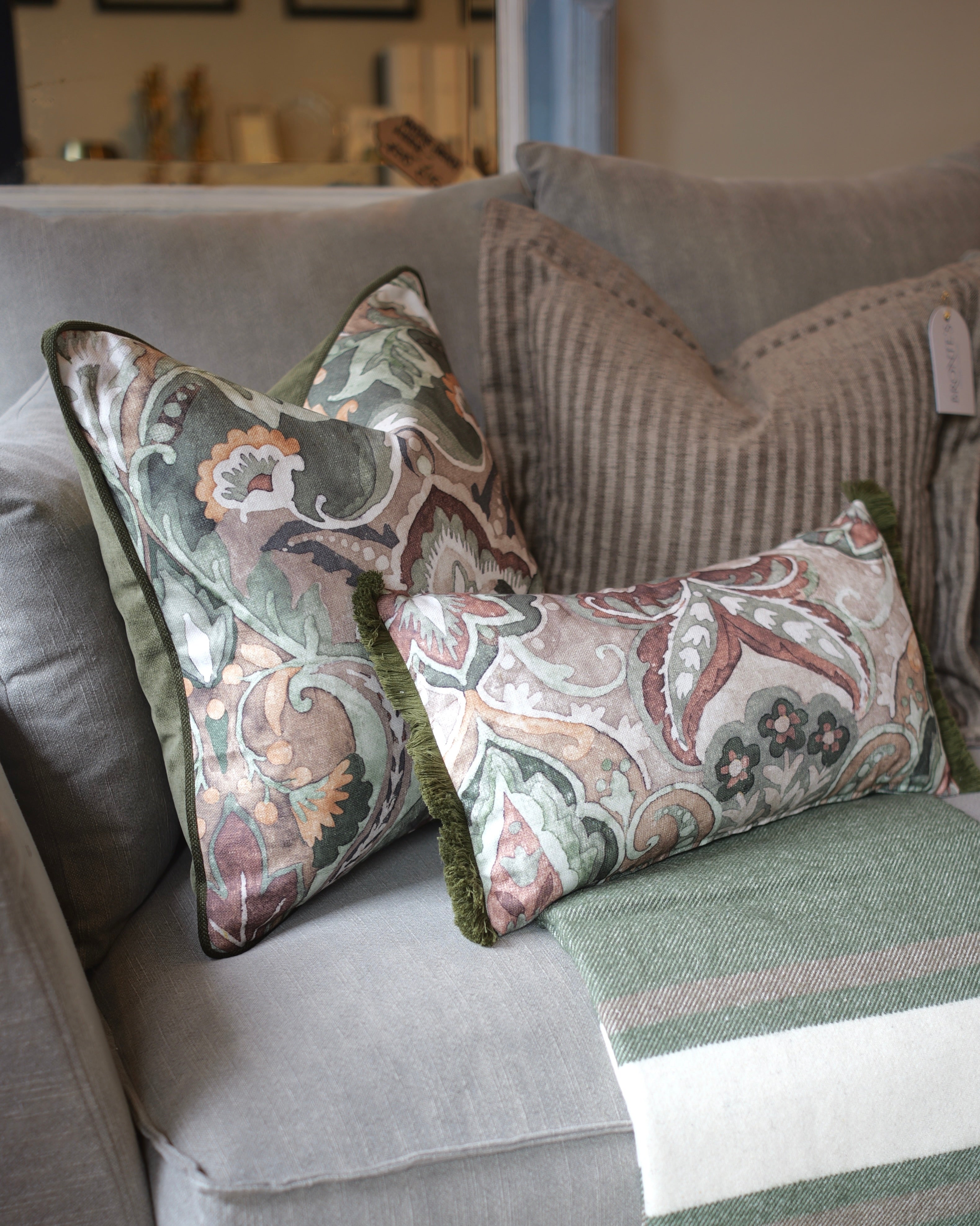 Two decorative throw pillows with a muted paisley pattern in sage green, dusty pink, and cream, styled on a gray sofa with a striped knit blanket.