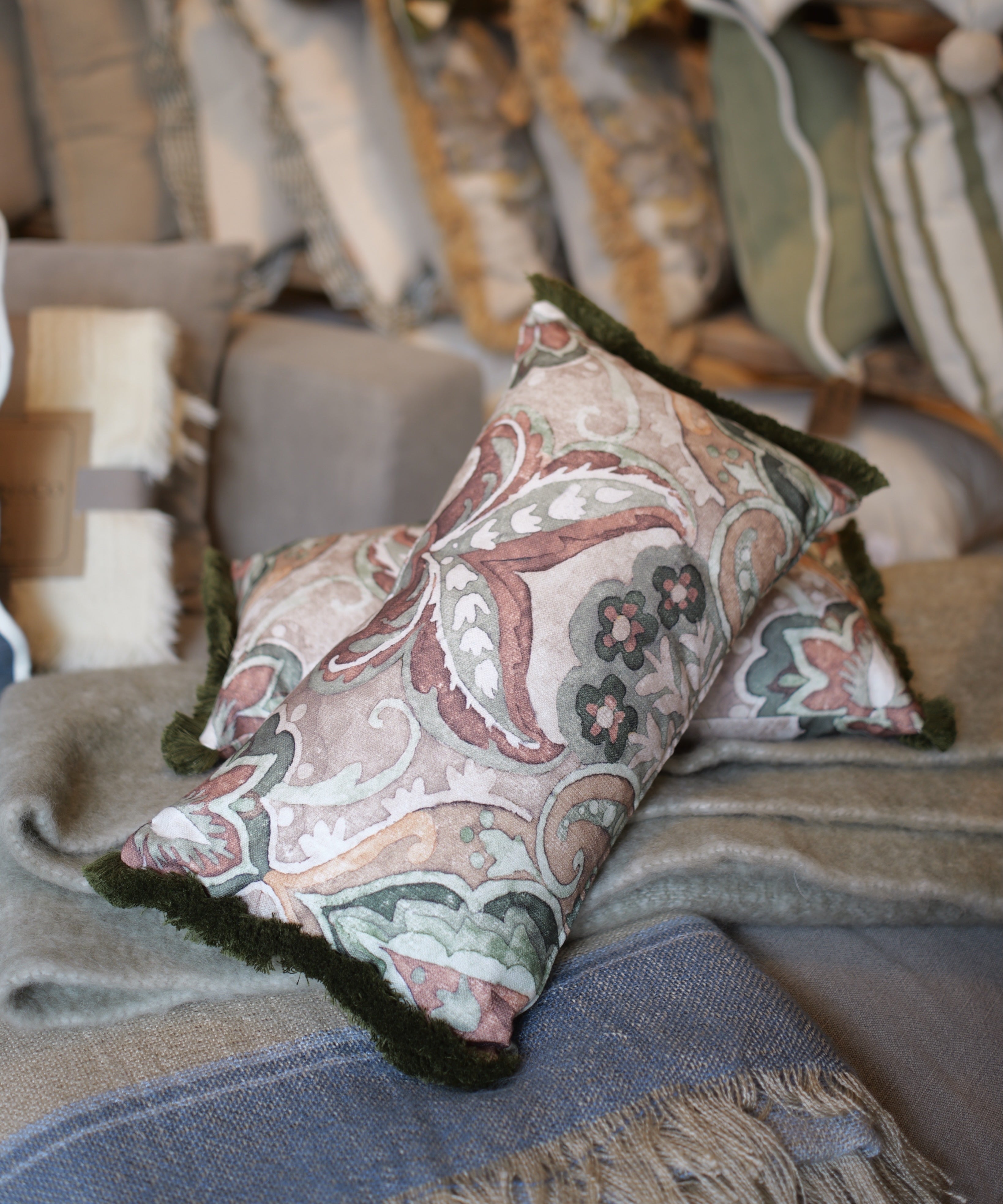 Two decorative throw pillows with a muted paisley pattern in dusty pink, sage green, and cream, accented with dark green fringed edges, styled on a gray blanket with other home textiles in the background.