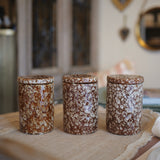 Three cylindrical ceramic canisters with matching lids, decorated in a brown and cream floral marbled pattern, arranged on a burlap runner in a softly blurred home interior.