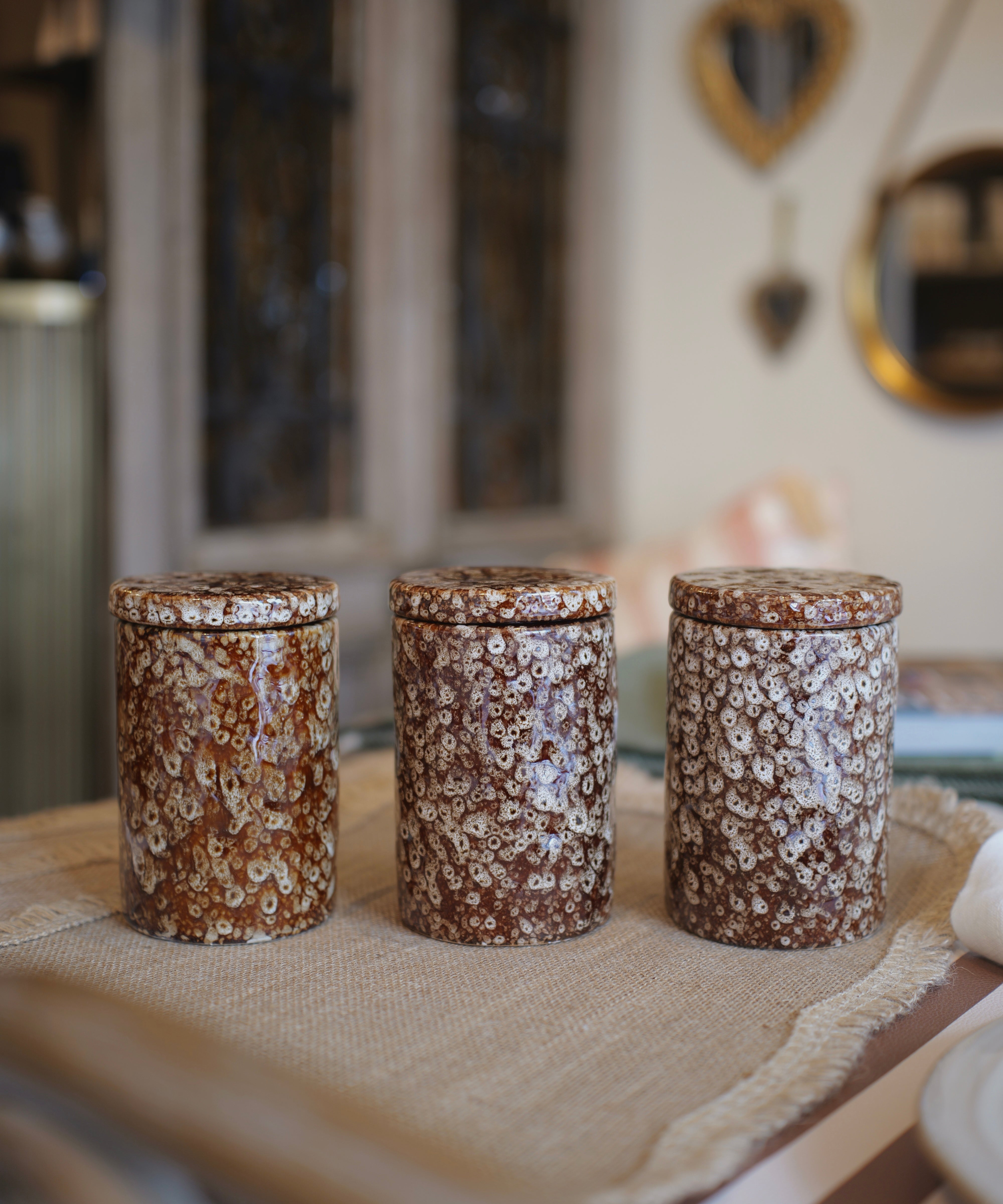 Three cylindrical ceramic canisters with matching lids, decorated in a brown and cream floral marbled pattern, arranged on a burlap runner in a softly blurred home interior.