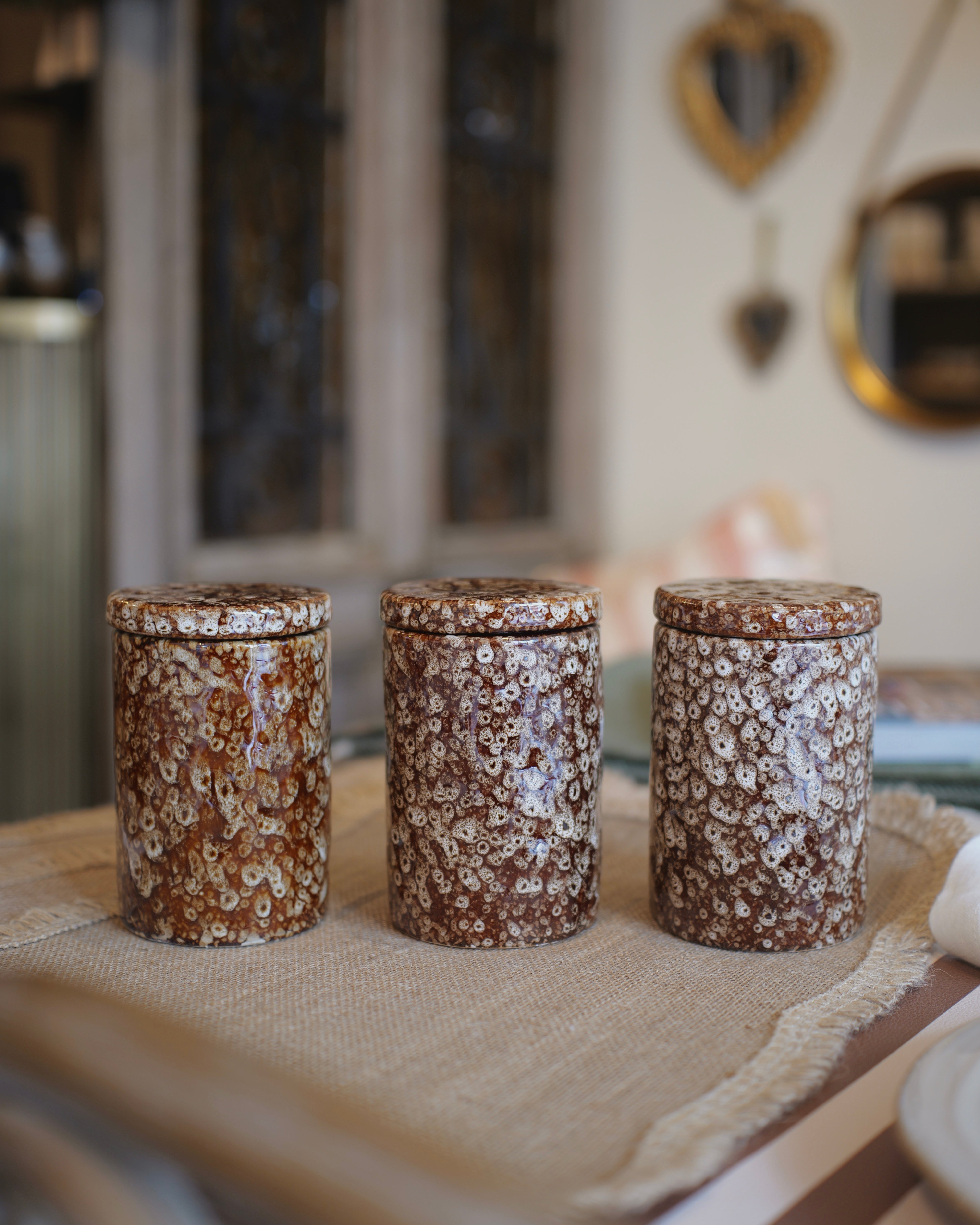 Three cylindrical ceramic canisters with matching lids, decorated in a brown and cream floral marbled pattern, arranged on a burlap runner in a softly blurred home interior.