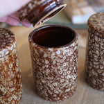Three brown and cream speckled ceramic canisters with lids are shown on a burlap surface, with a hand lifting the lid of the center canister.