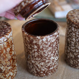 Three brown and cream speckled ceramic canisters with lids are shown on a burlap surface, with a hand lifting the lid of the center canister.