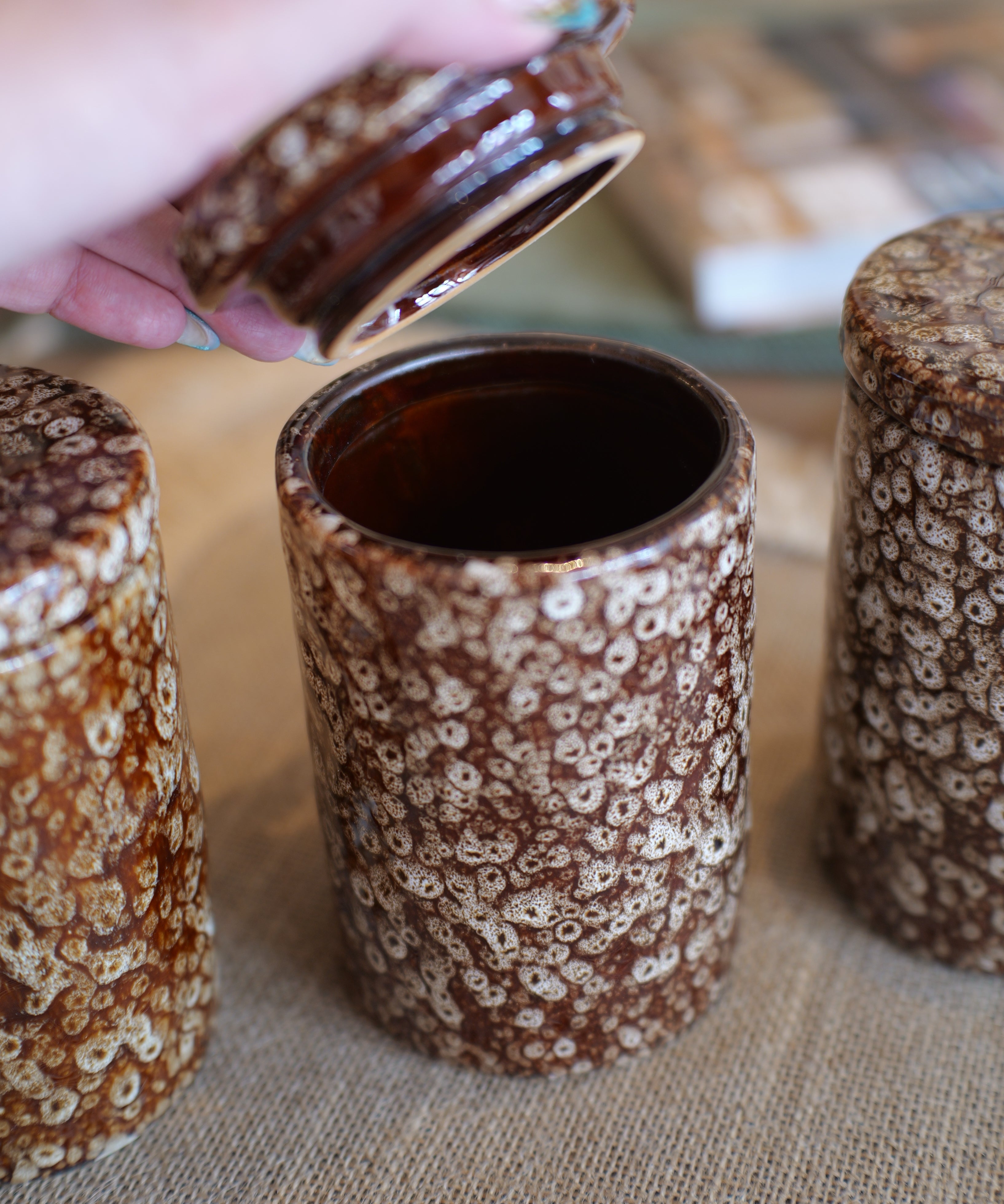 Three brown and cream speckled ceramic canisters with lids are shown on a burlap surface, with a hand lifting the lid of the center canister.