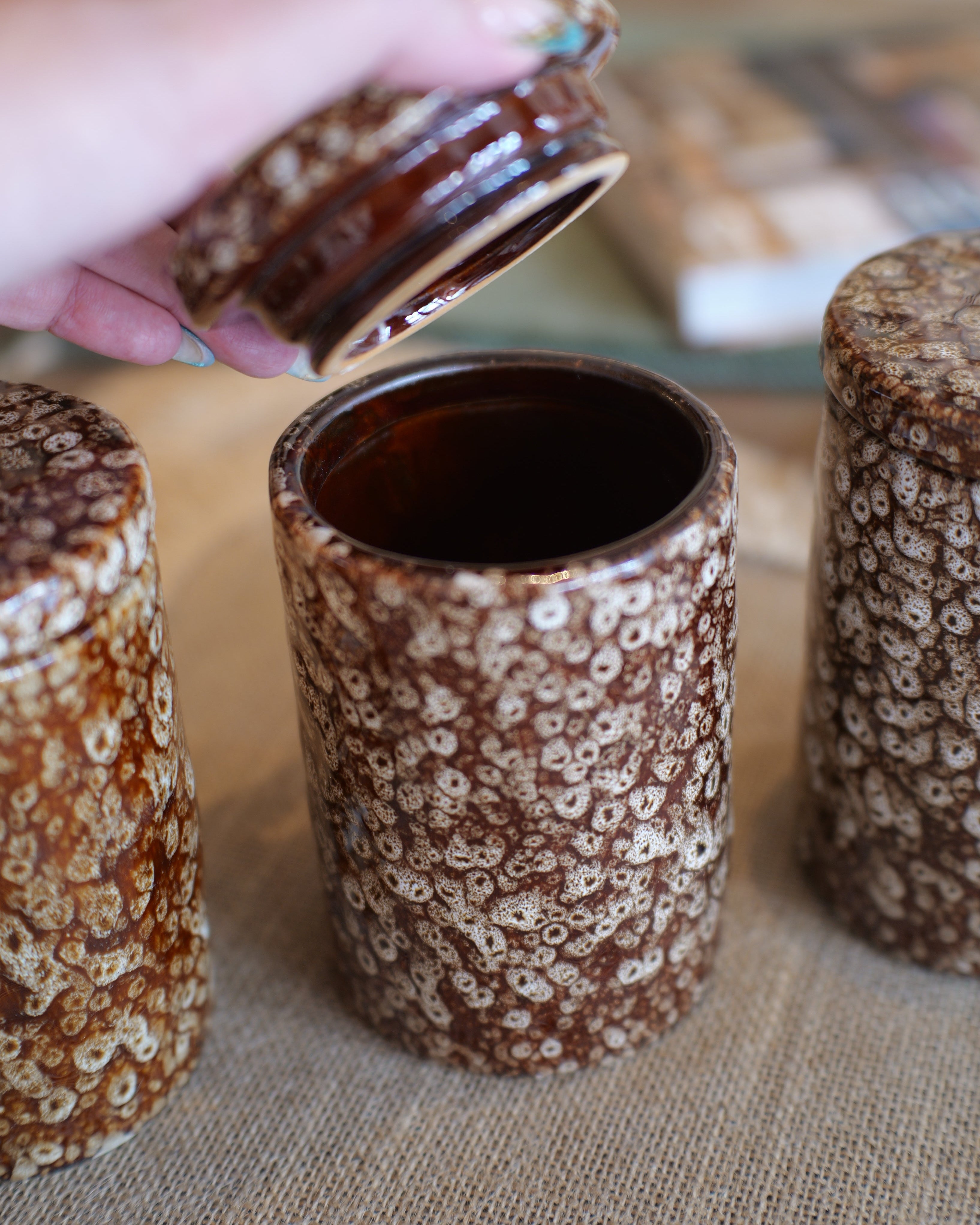 Three brown and cream speckled ceramic canisters with lids are shown on a burlap surface, with a hand lifting the lid of the center canister.