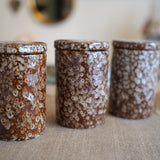 Three small cylindrical lidded jars with a brown and cream floral marbled pattern, arranged on a beige fabric surface with a softly blurred indoor background.