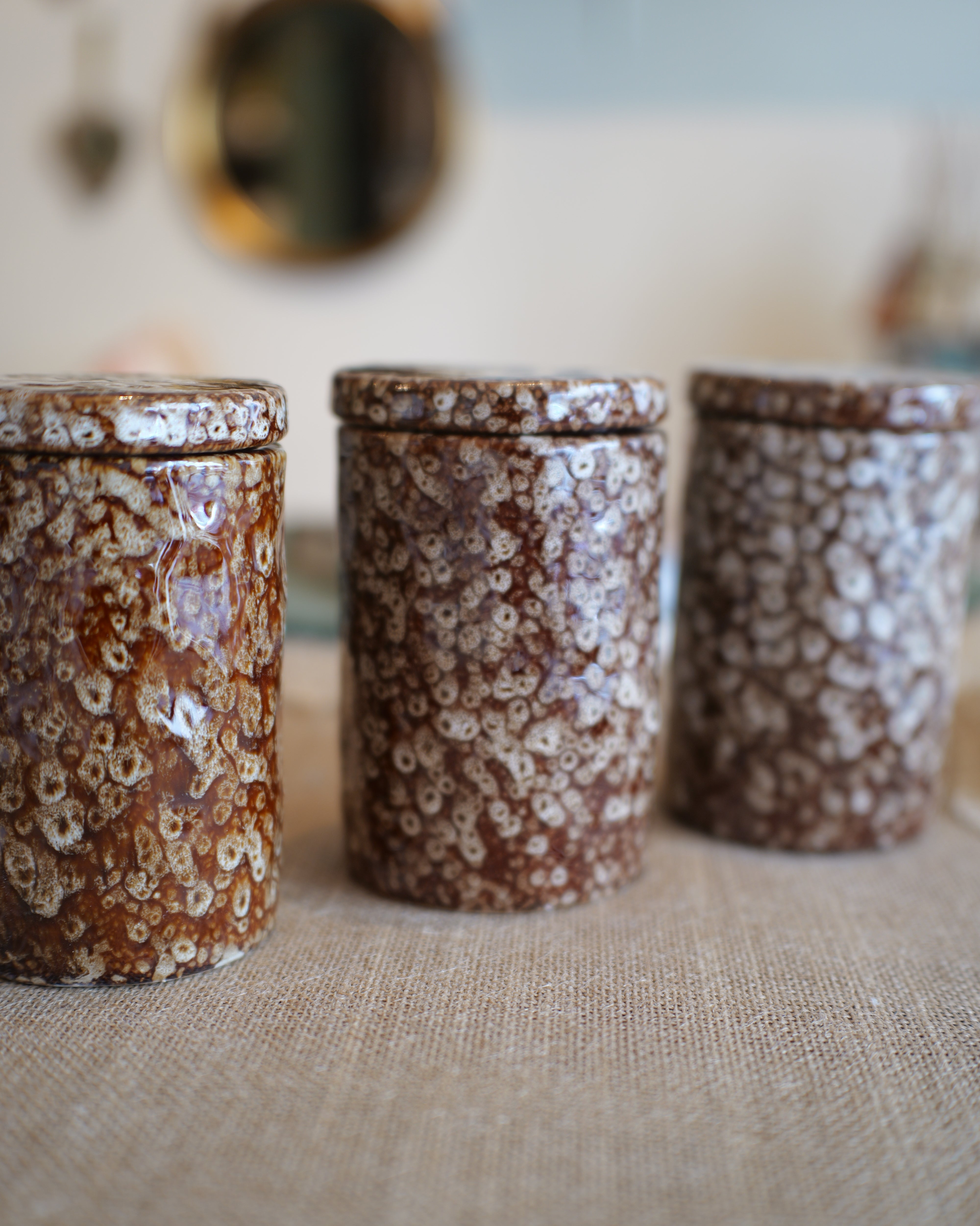 Three small cylindrical lidded jars with a brown and cream floral marbled pattern, arranged on a beige fabric surface with a softly blurred indoor background.