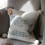 A square decorative throw pillow with pom-poms and a printed hare motif, styled on a gray sofa in soft natural light.