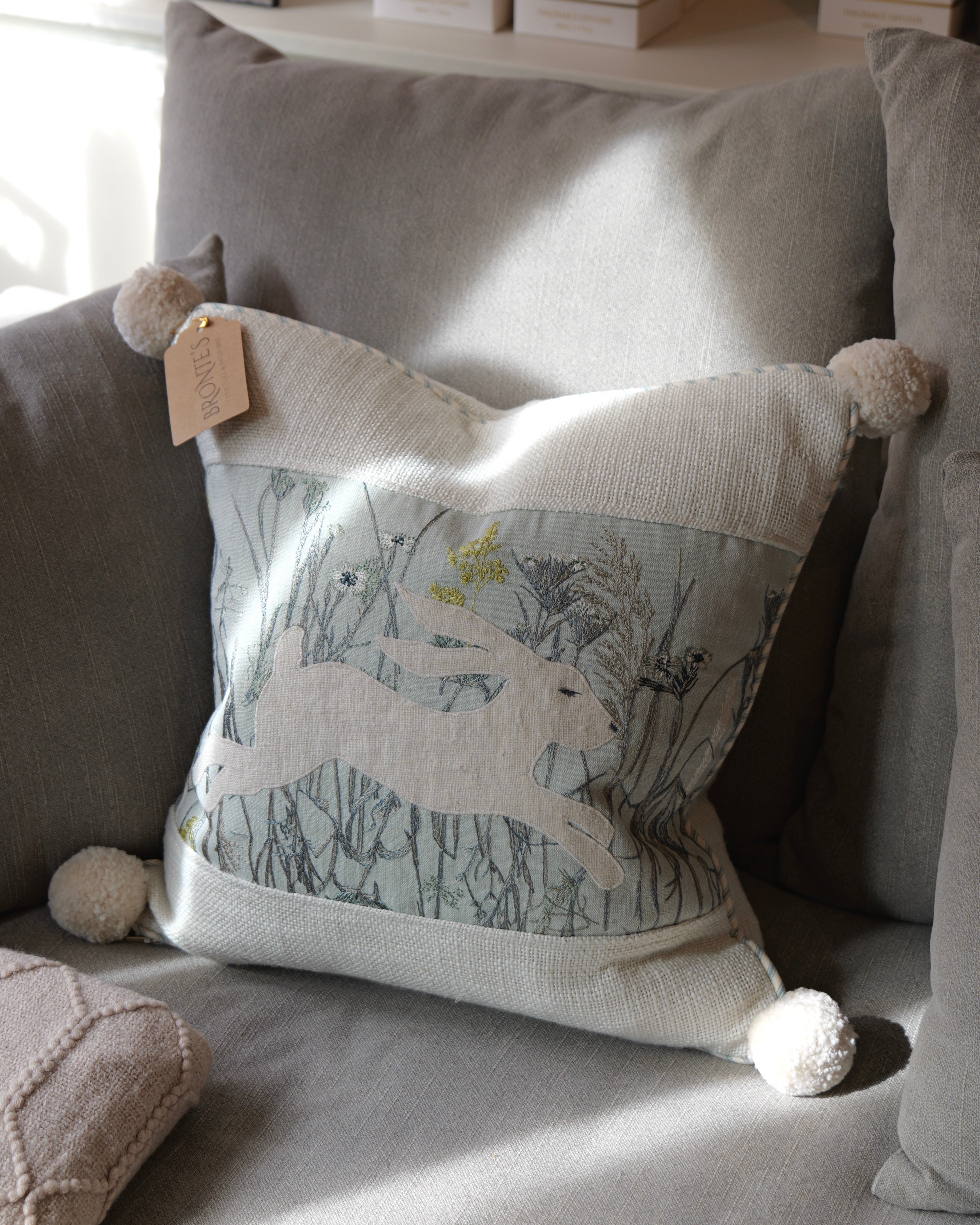 A square decorative throw pillow with pom-poms and a printed hare motif, styled on a gray sofa in soft natural light.