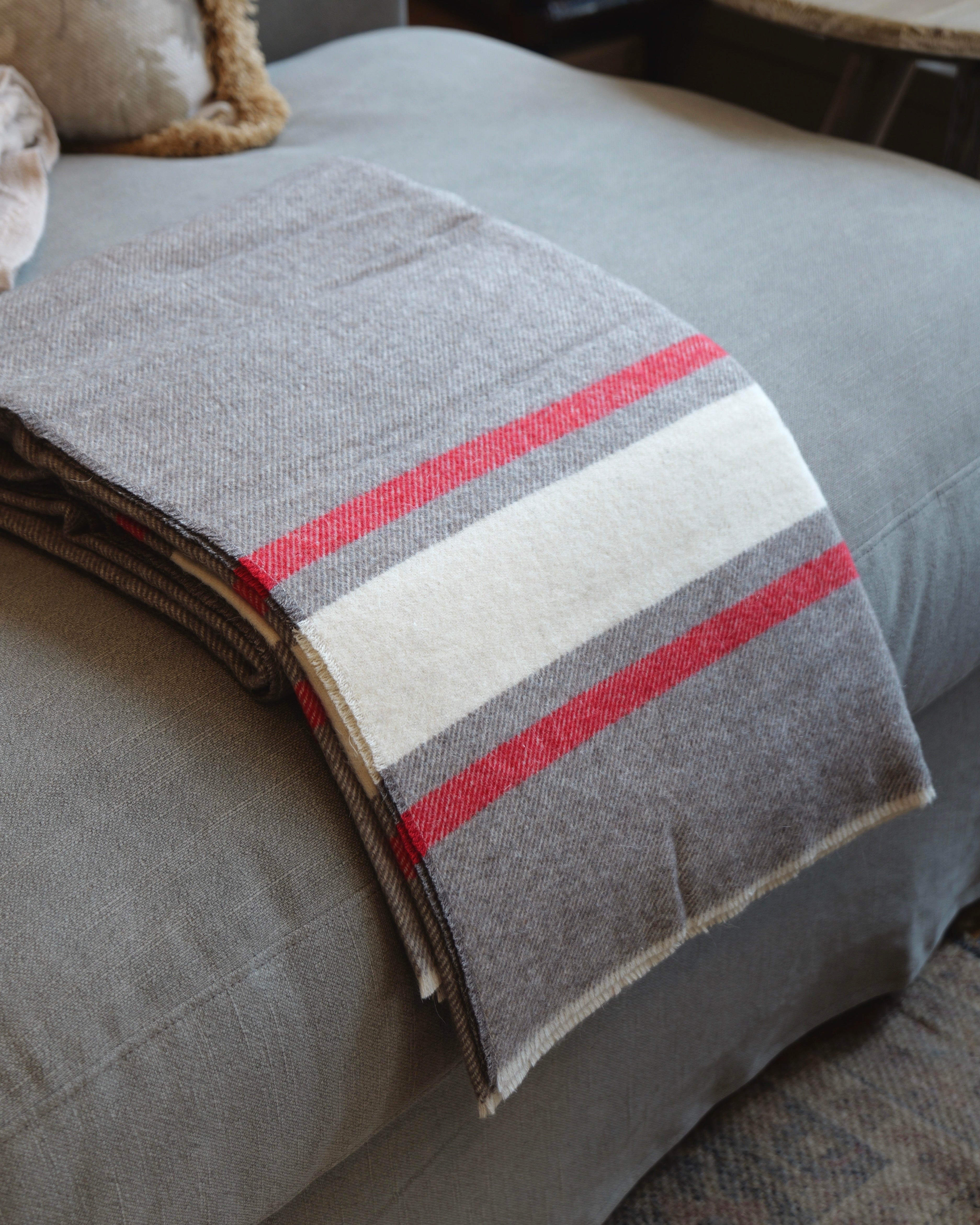 A folded wool blanket with a gray, white, and red stripe pattern draped over a gray upholstered chair in a home setting.