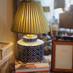 Decorative lamp with pleated shade on a stack of books, surrounded by wicker furniture and a framed picture.