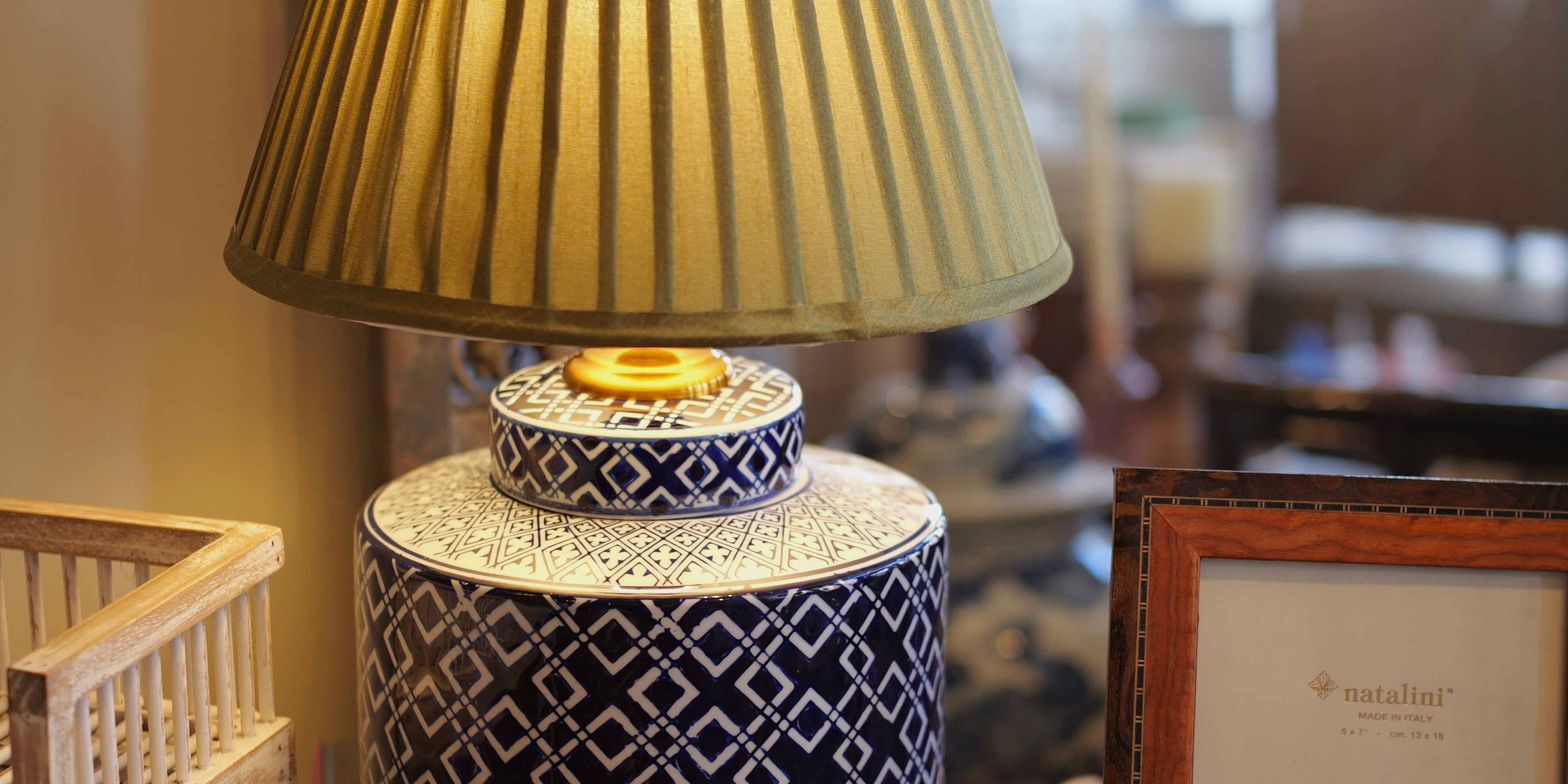 Decorative lamp with pleated shade on a stack of books, surrounded by wicker furniture and a framed picture.