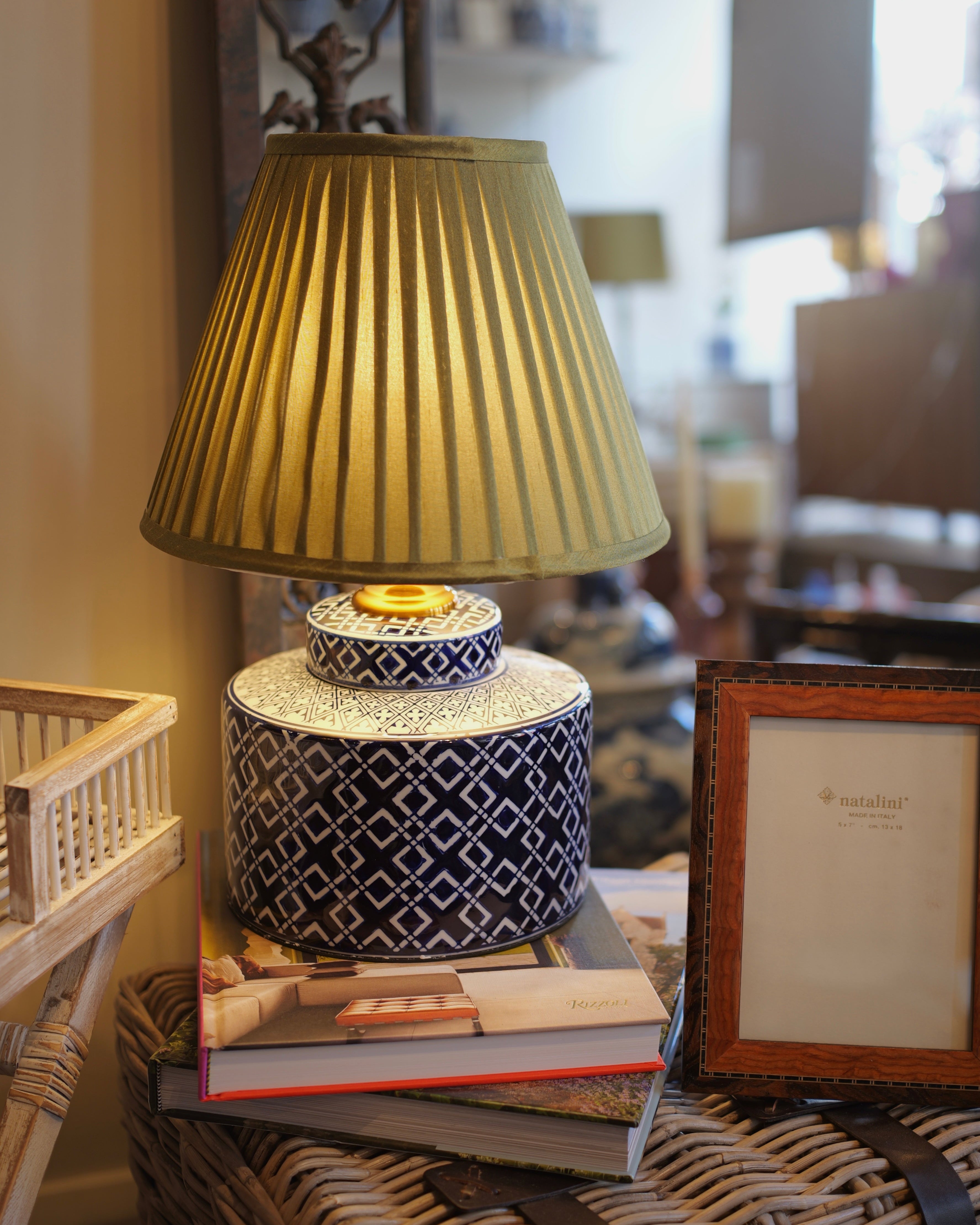 Decorative lamp with pleated shade on a stack of books, surrounded by wicker furniture and a framed picture.