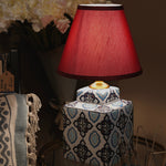 Decorative table lamp with a red shade on a patterned base, surrounded by flowers and decor items.