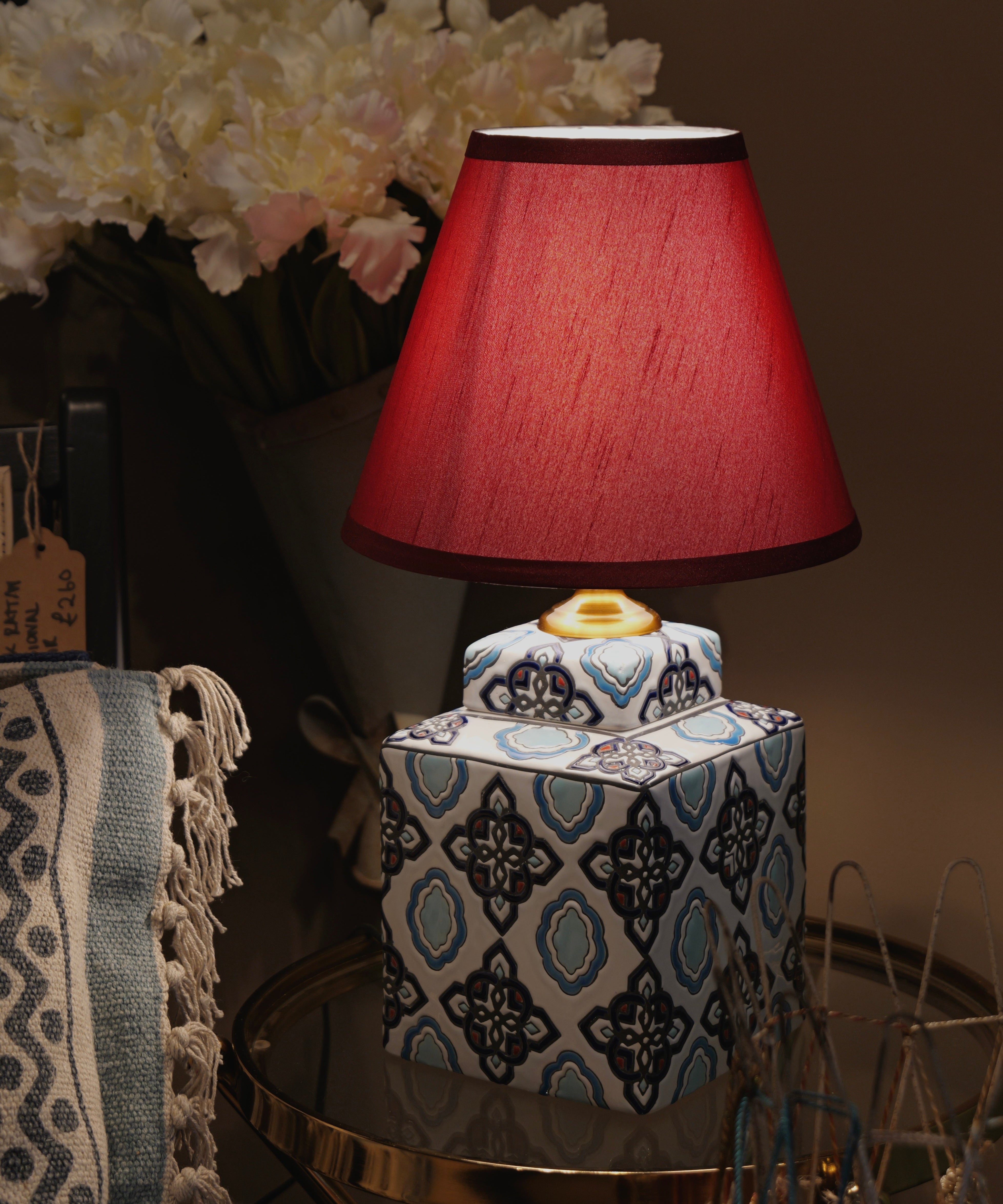 Decorative table lamp with a red shade on a patterned base, surrounded by flowers and decor items.
