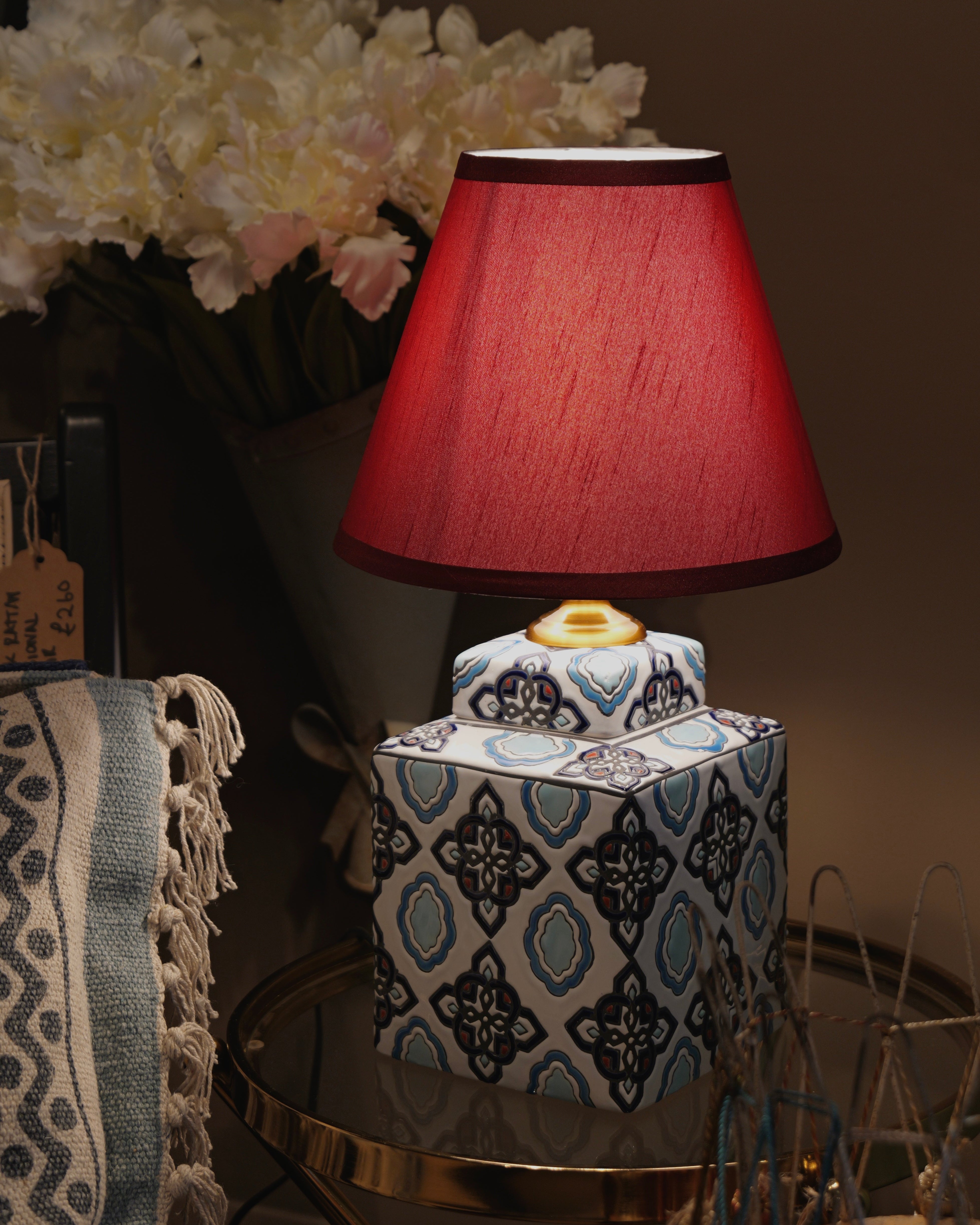 Decorative table lamp with a red shade on a patterned base, surrounded by flowers and decor items.