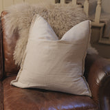 Light gray pillow on a brown leather couch with a fluffy blanket in the background.