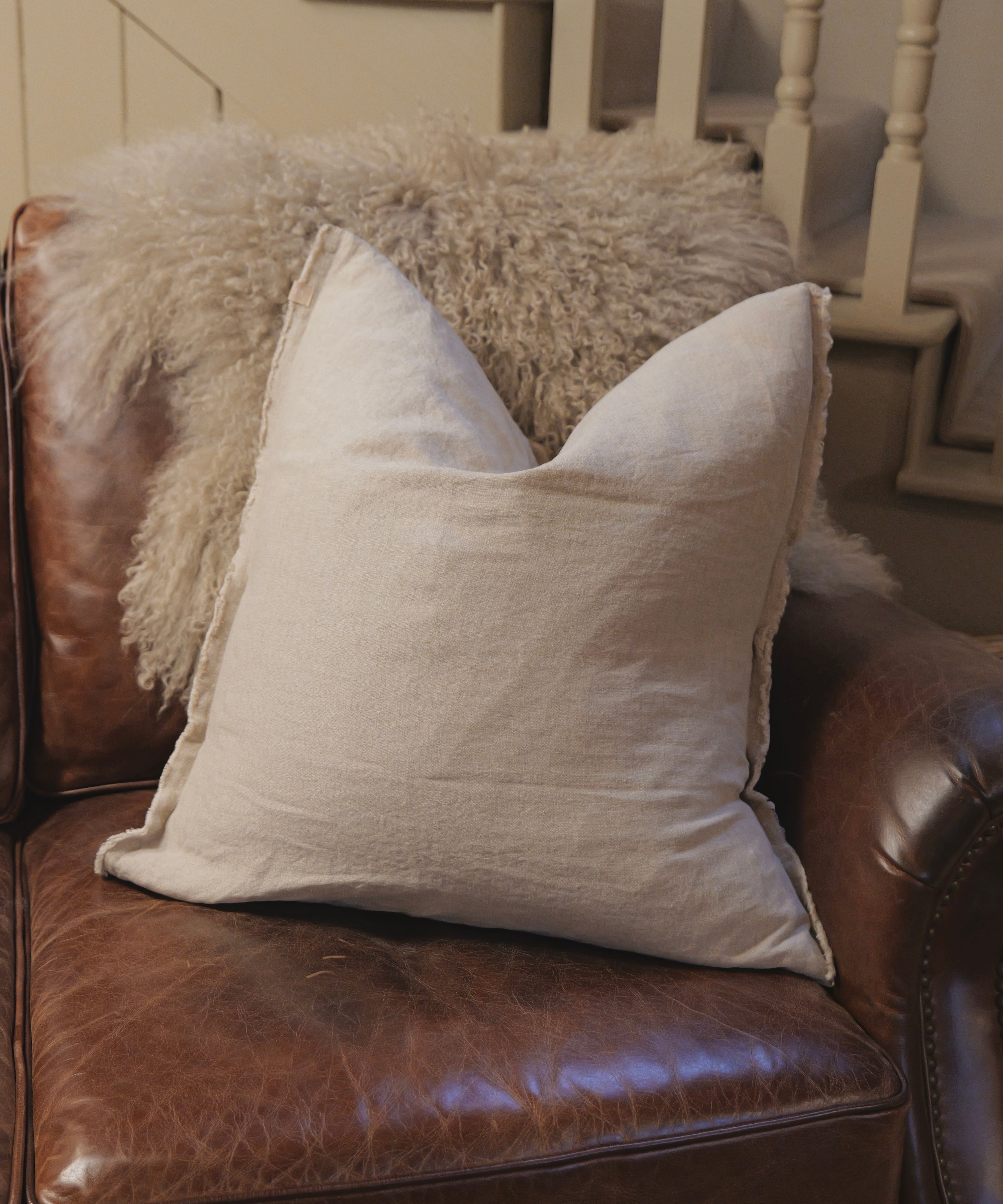 Light gray pillow on a brown leather couch with a fluffy blanket in the background.