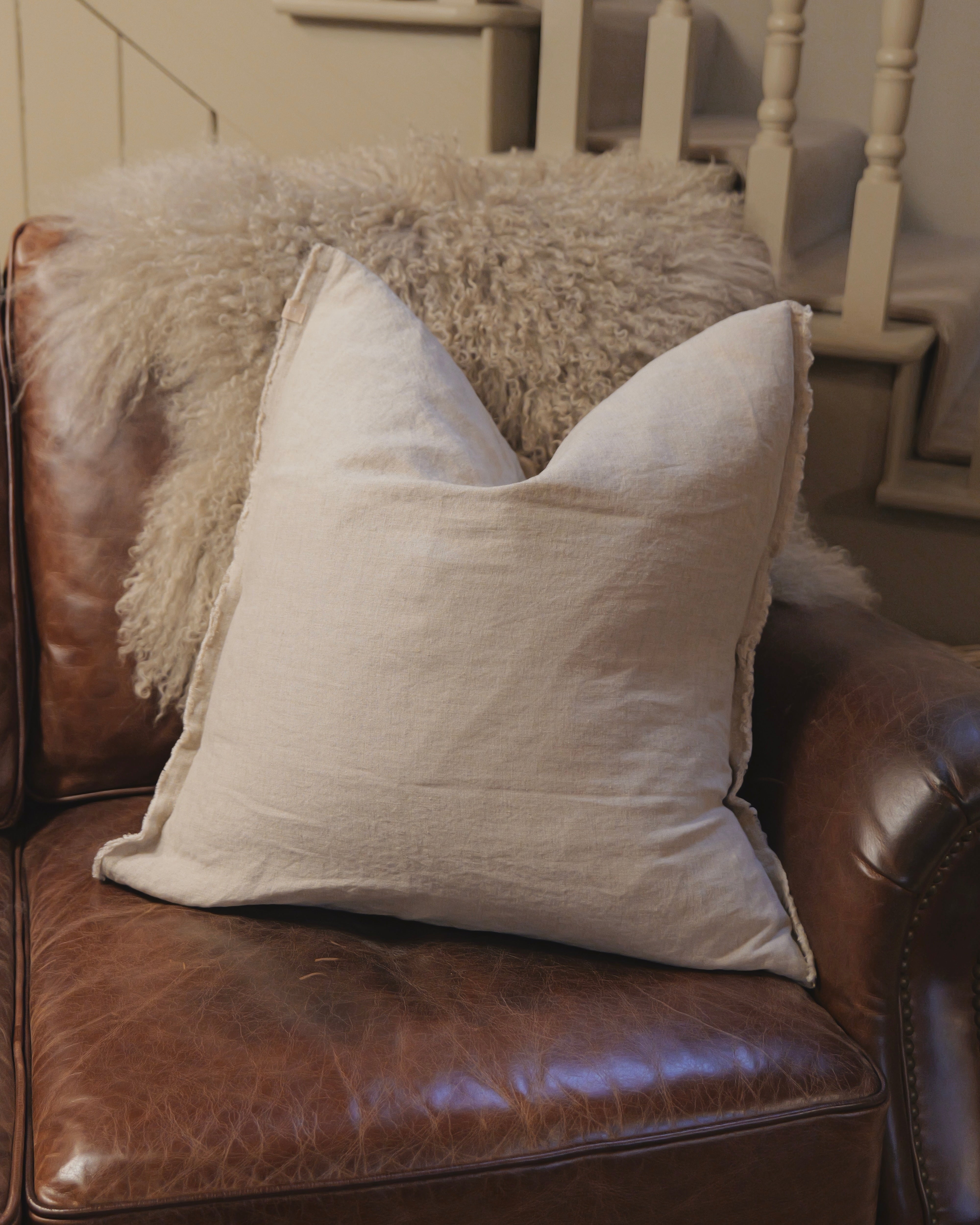Light gray pillow on a brown leather couch with a fluffy blanket in the background.